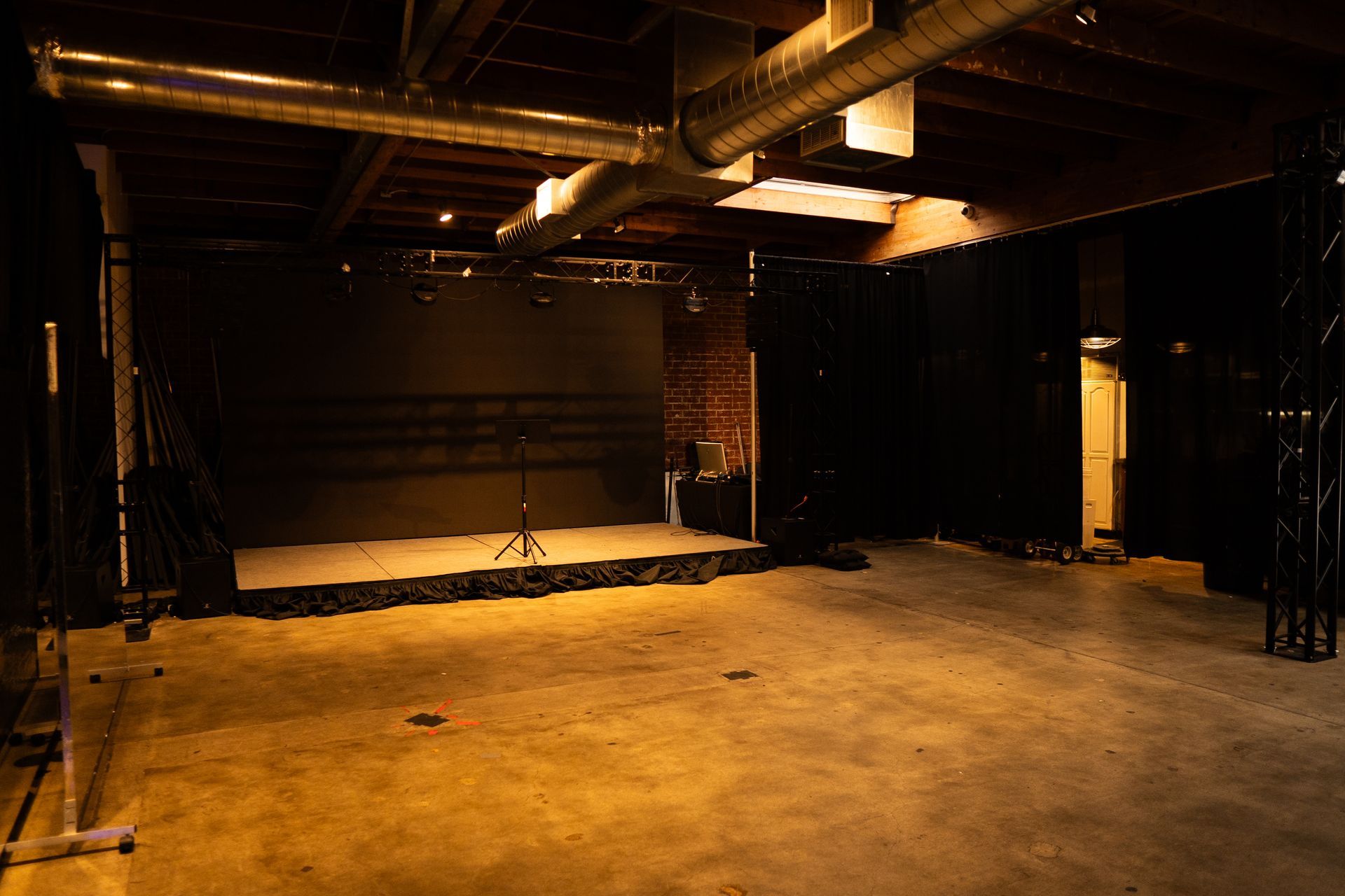 A dimly lit event space featuring a small, raised stage with a microphone stand against a dark backdrop and exposed beams.