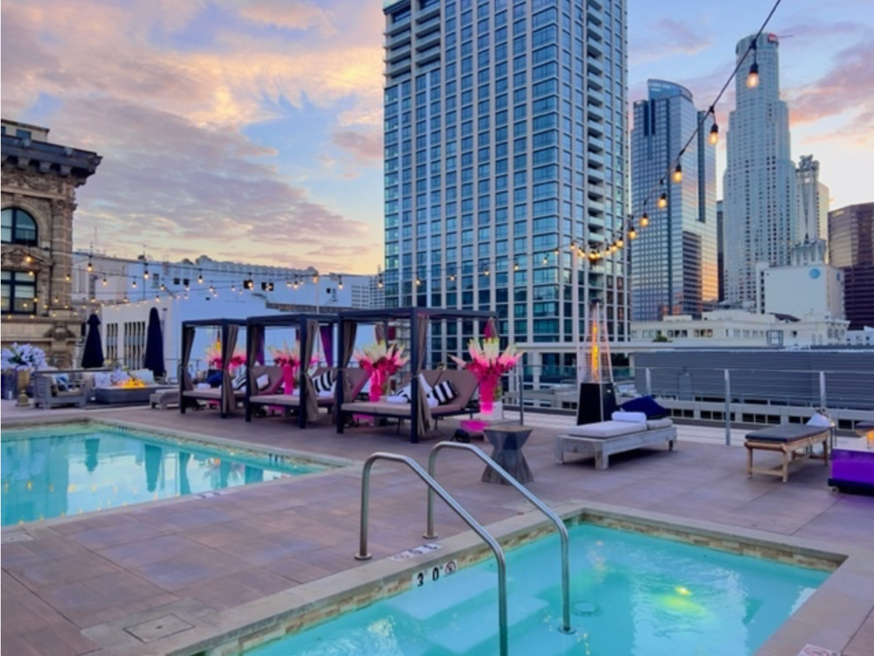 A rooftop pool and hot tub area at sunset with lounge chairs, cabanas, and tall city buildings in the background.