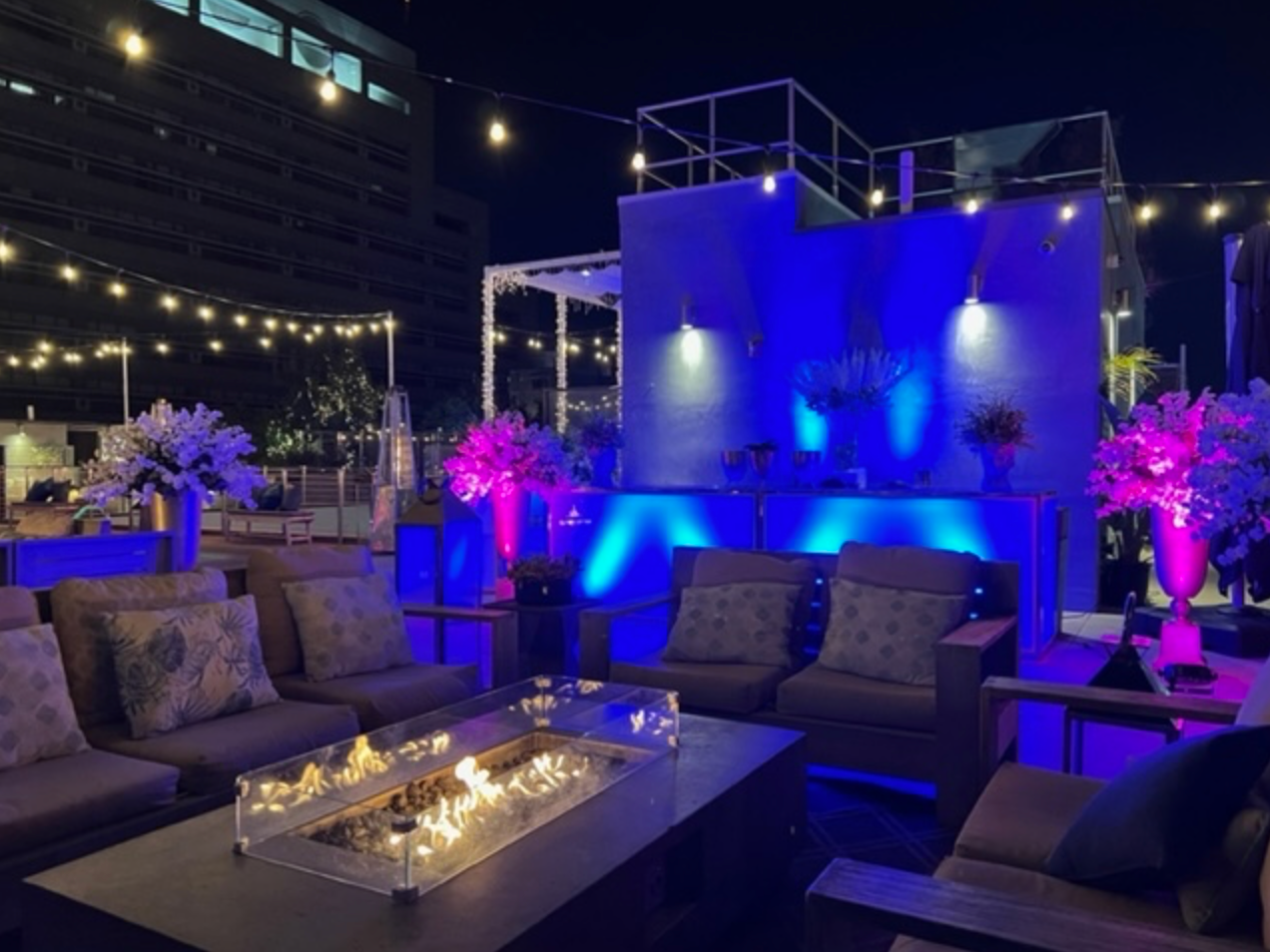 An outdoor rooftop lounge at night featuring gray sofas around a fire pit, illuminated by blue lights and string lights.