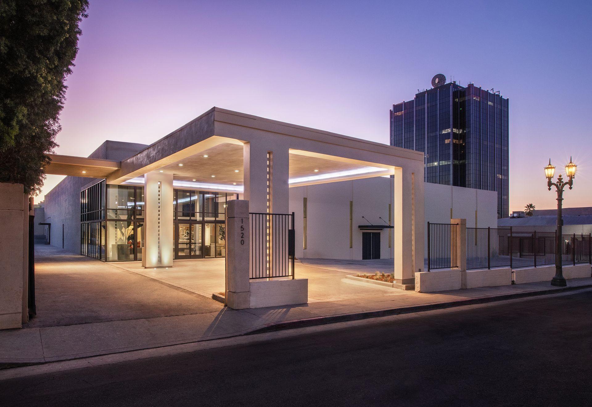 Modern white building entrance with a covered driveway, illuminated at dusk with a skyscraper in the background.