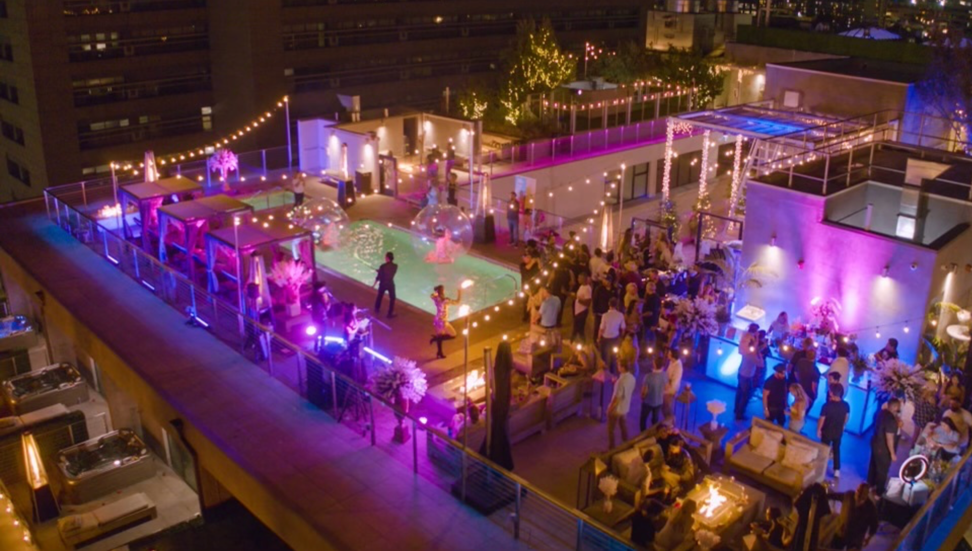 People gather on a rooftop lounge at night, featuring a lit swimming pool, string lights, and purple ambient lighting.