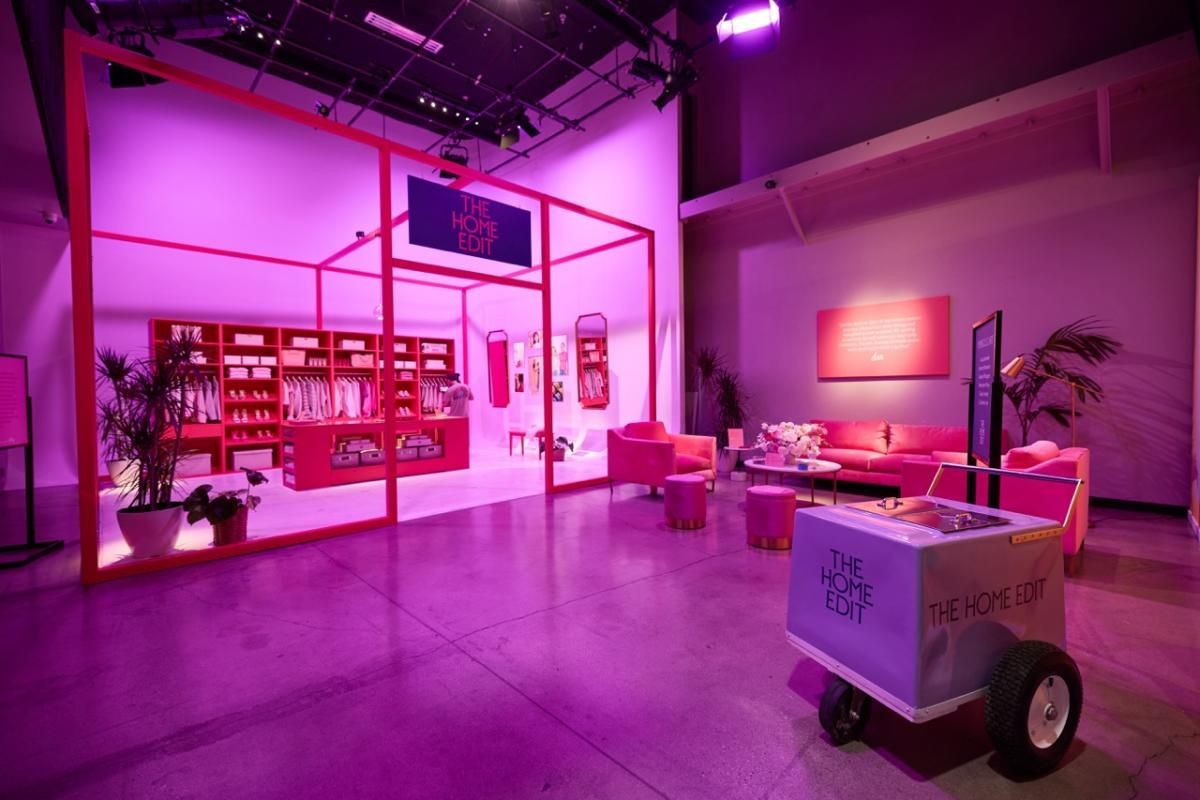 A bright, magenta-lit indoor event space featuring a product display booth, lounge seating, and a branded rolling cart.