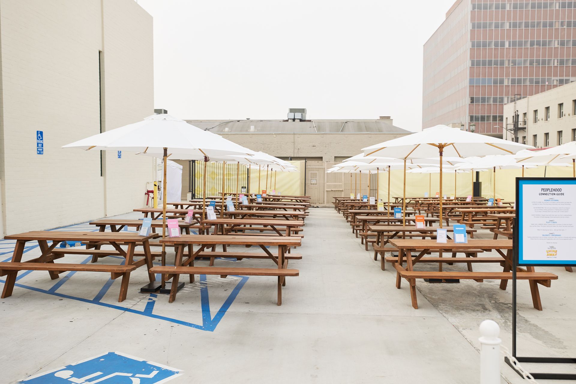 Outdoor dining area in a parking lot with rows of wooden picnic tables, white umbrellas, and blue handicap markings.