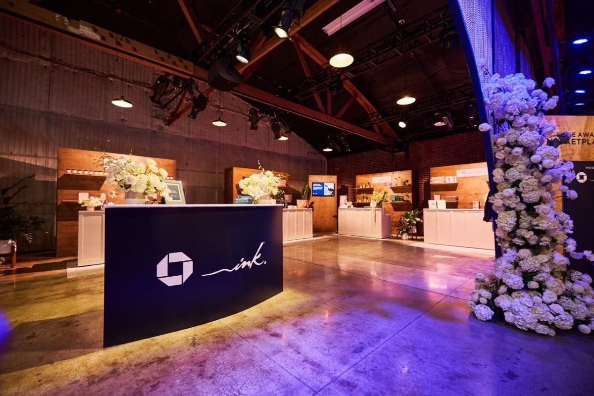A modern event venue interior featuring a dark reception desk with a white logo, wooden shelving, and a floral pillar.