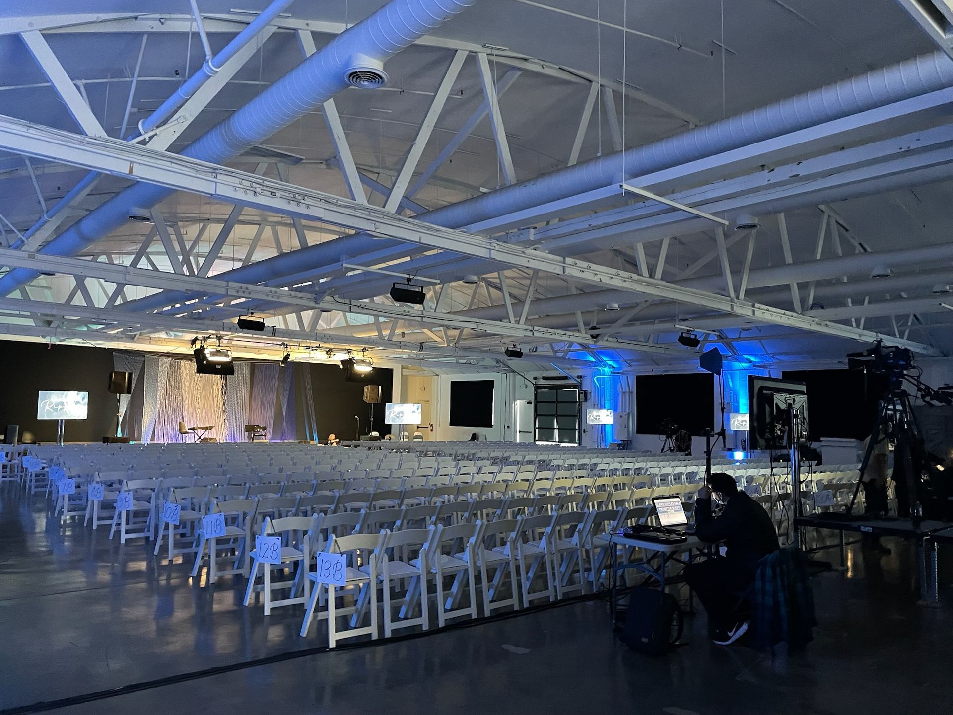 A large event space with rows of white chairs facing a stage with light blue ambient lighting and a technical setup.