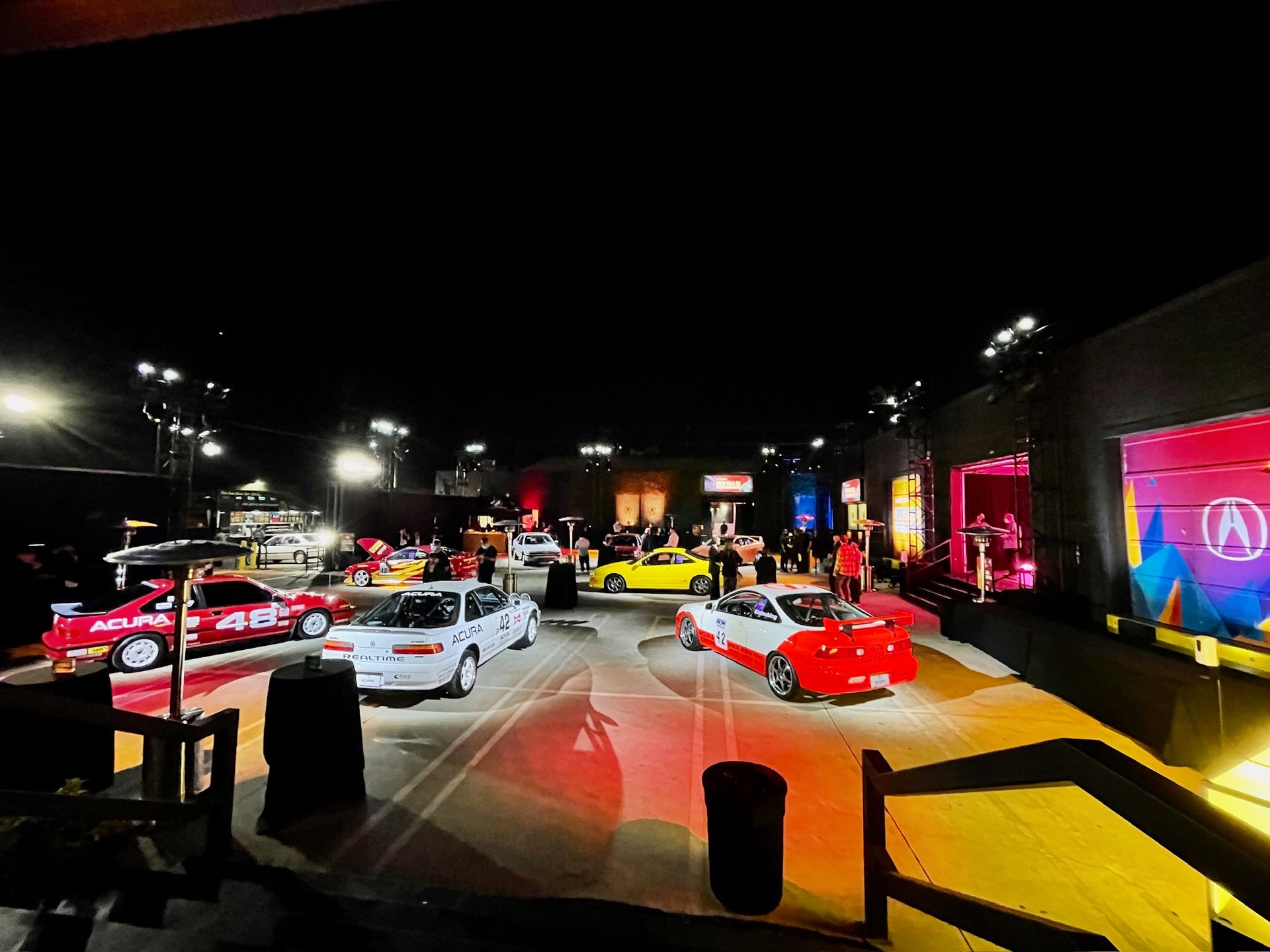 Several sports cars displayed in an outdoor evening venue with warm, dramatic lighting.