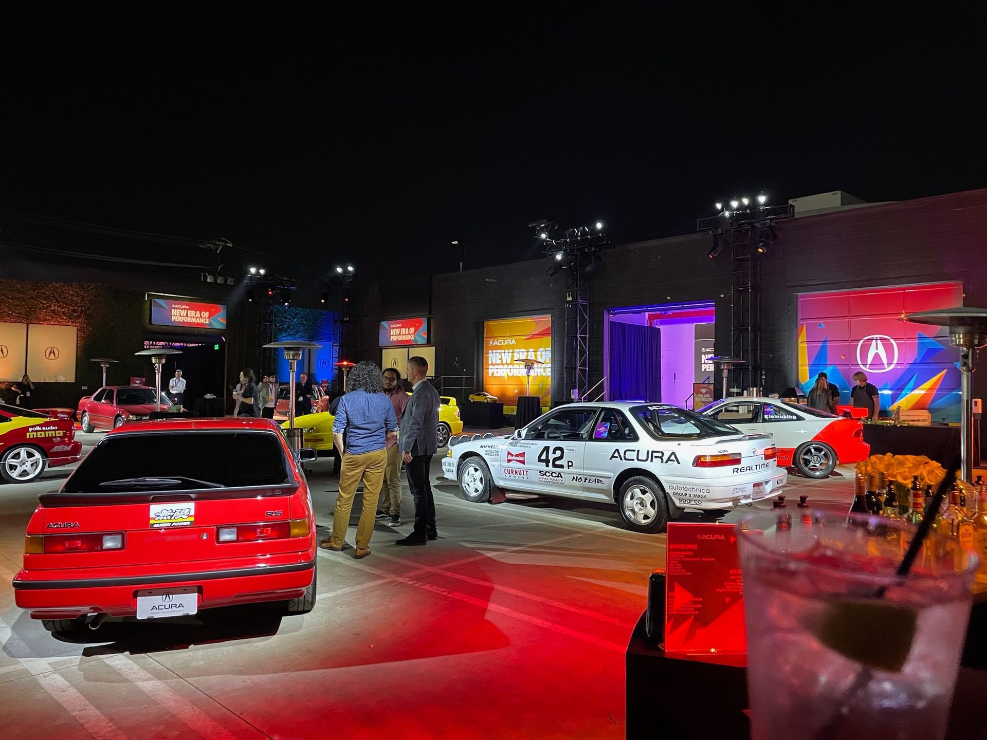 A car show at night features a red Acura, a white racing Acura with number 42, and event attendees in a paved lot.