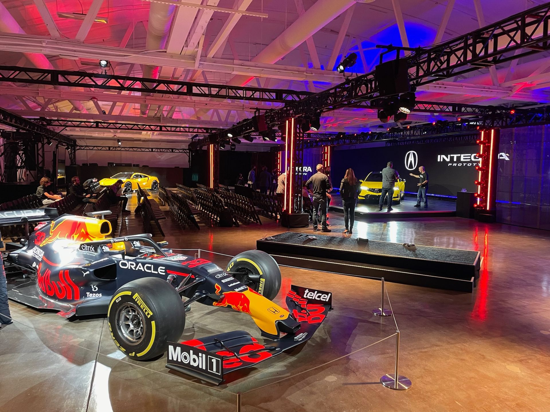 A Red Bull Formula 1 car on display in a room with event lighting and an Acura car on a stage in the background.