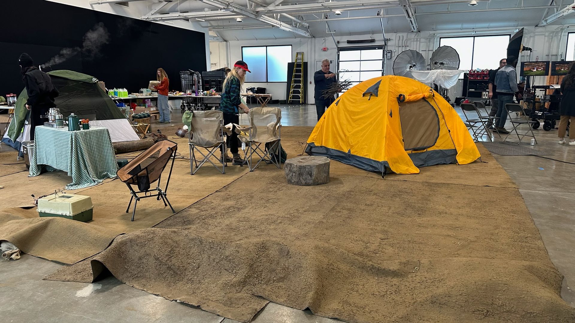 A photo studio setup features a yellow dome tent on a dirt-covered floor, with chairs, a table, and camping equipment nearby.