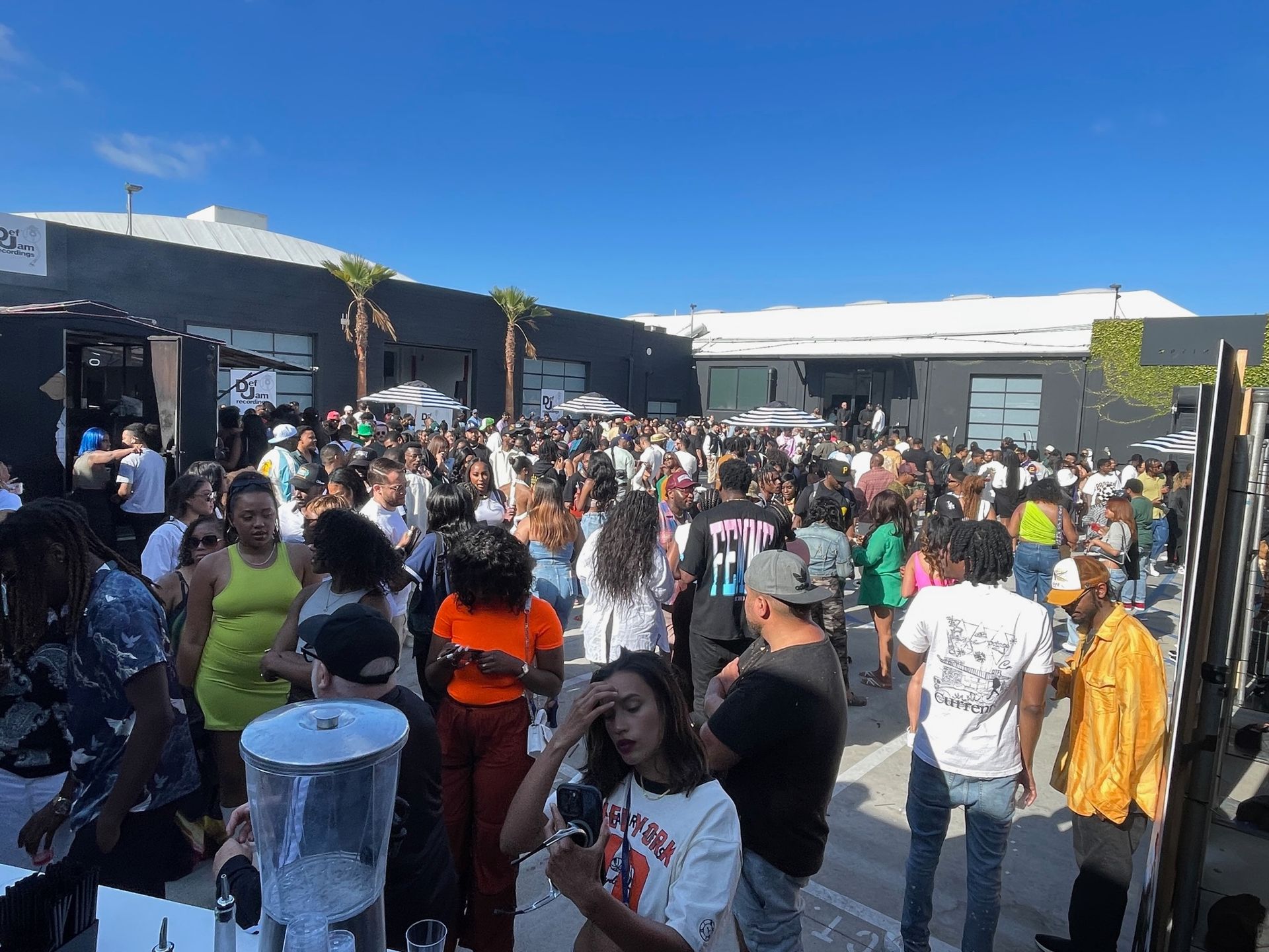 A large, outdoor crowd gathers for an event in a paved plaza between two modern, dark-colored warehouse buildings.