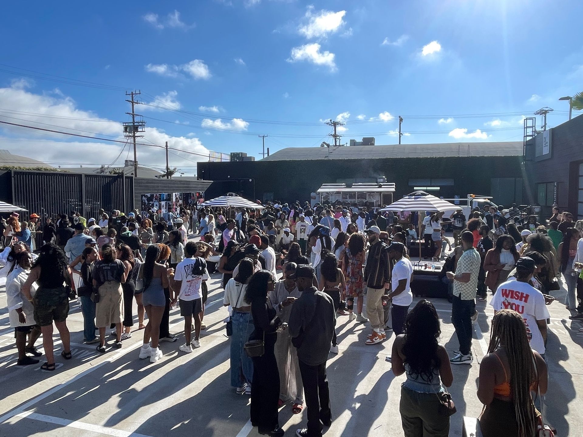 A large, diverse crowd gathers outdoors on a sunny day in an open concrete lot with buildings and umbrellas in view.