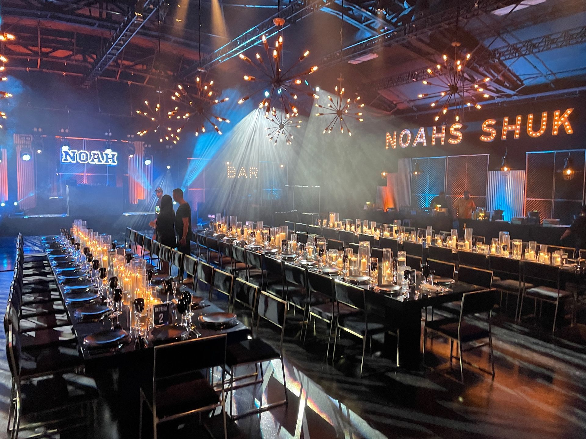 A dimly lit event space with long, banquet-style dining tables set for a formal gathering under festive light fixtures.