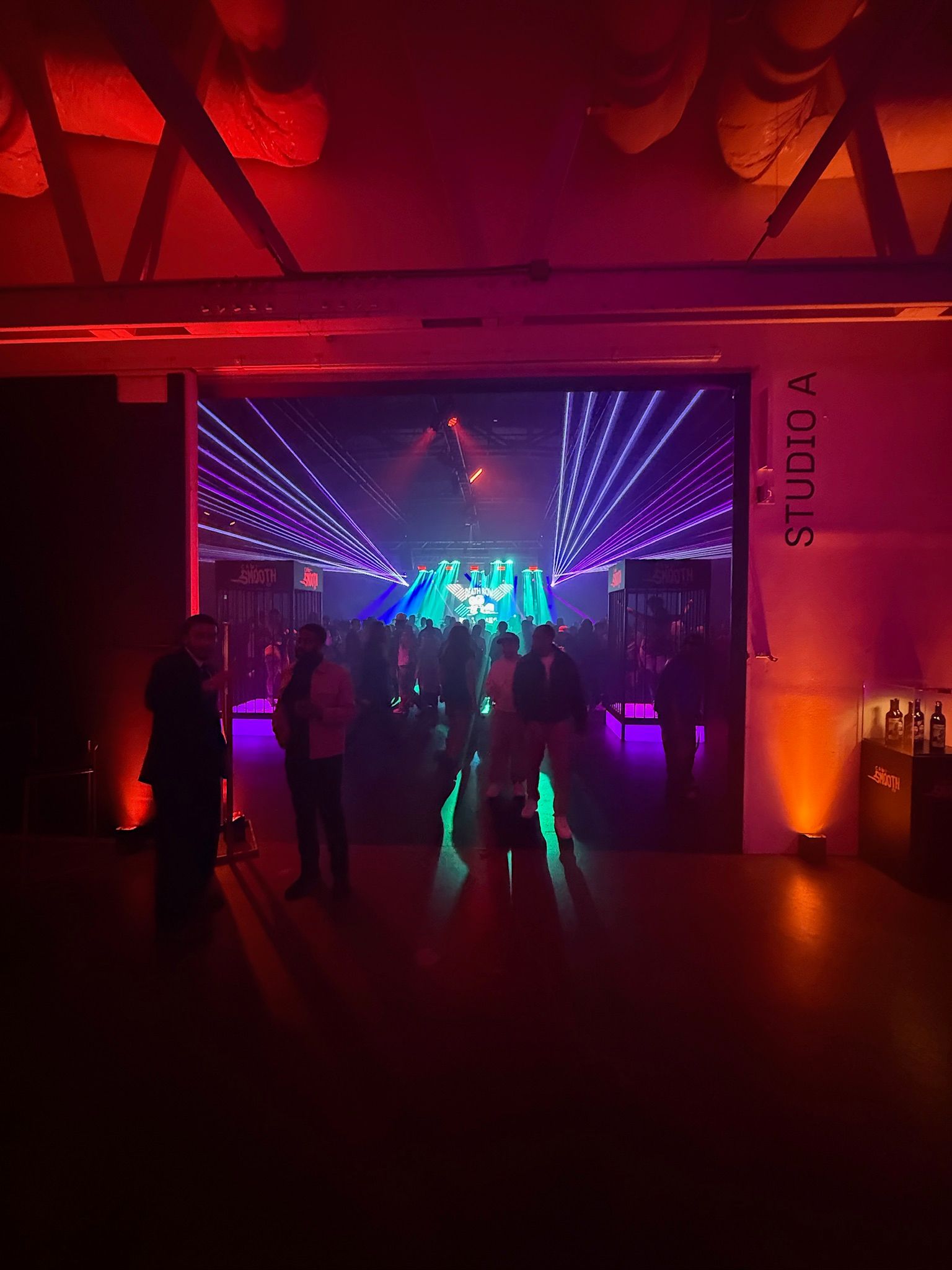 People gather in a dim, red-lit studio space, looking toward a bright, energetic performance stage with laser lighting.