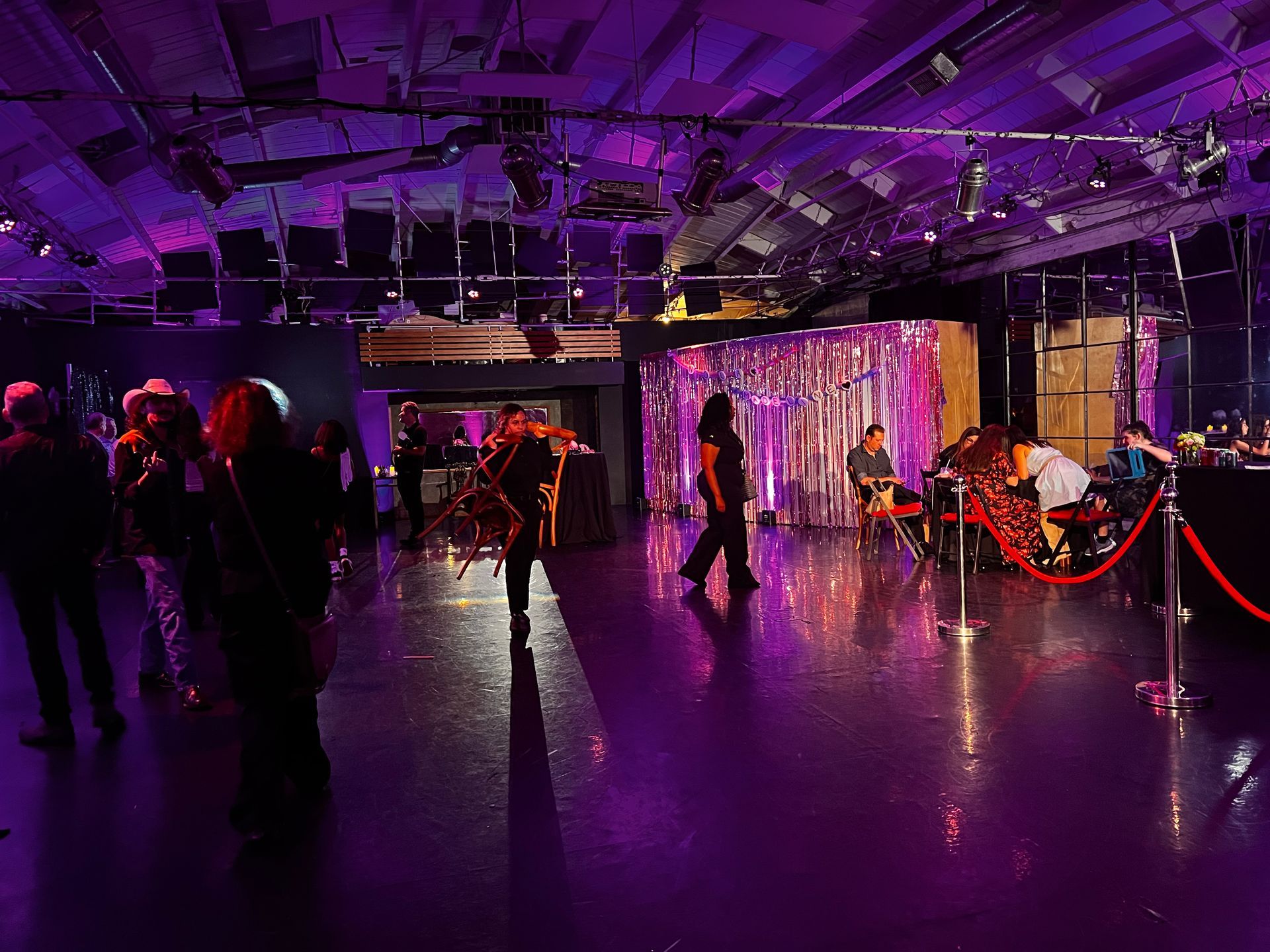 People mingle in a dark room bathed in purple light, featuring a shiny floor and shimmering metallic fringe curtains.