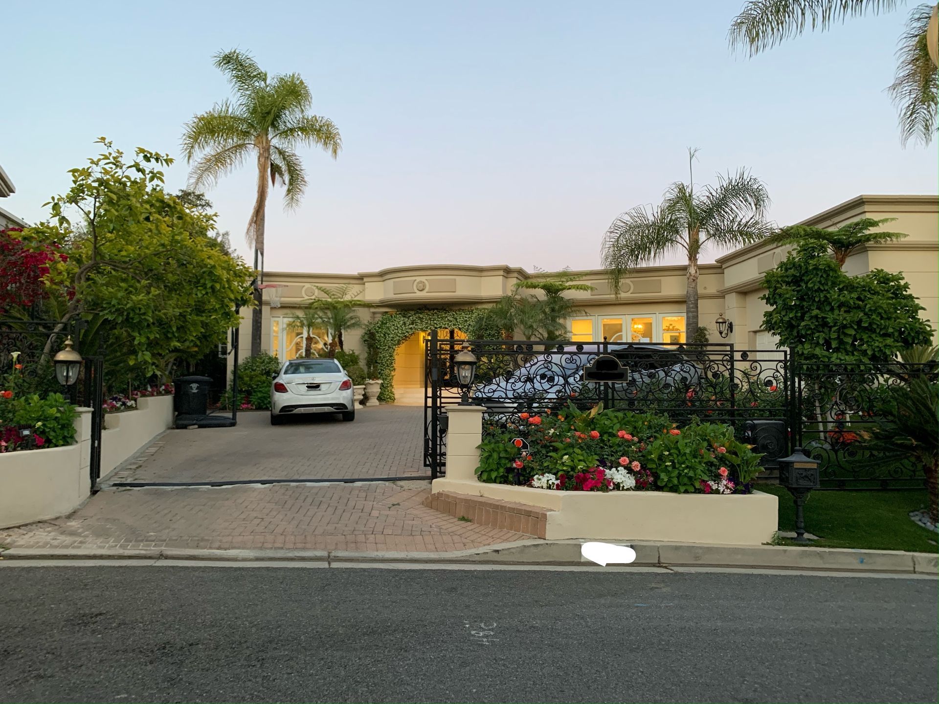 A beige, single-story house behind a black wrought-iron fence and gate, with palm trees and a car parked in the driveway.
