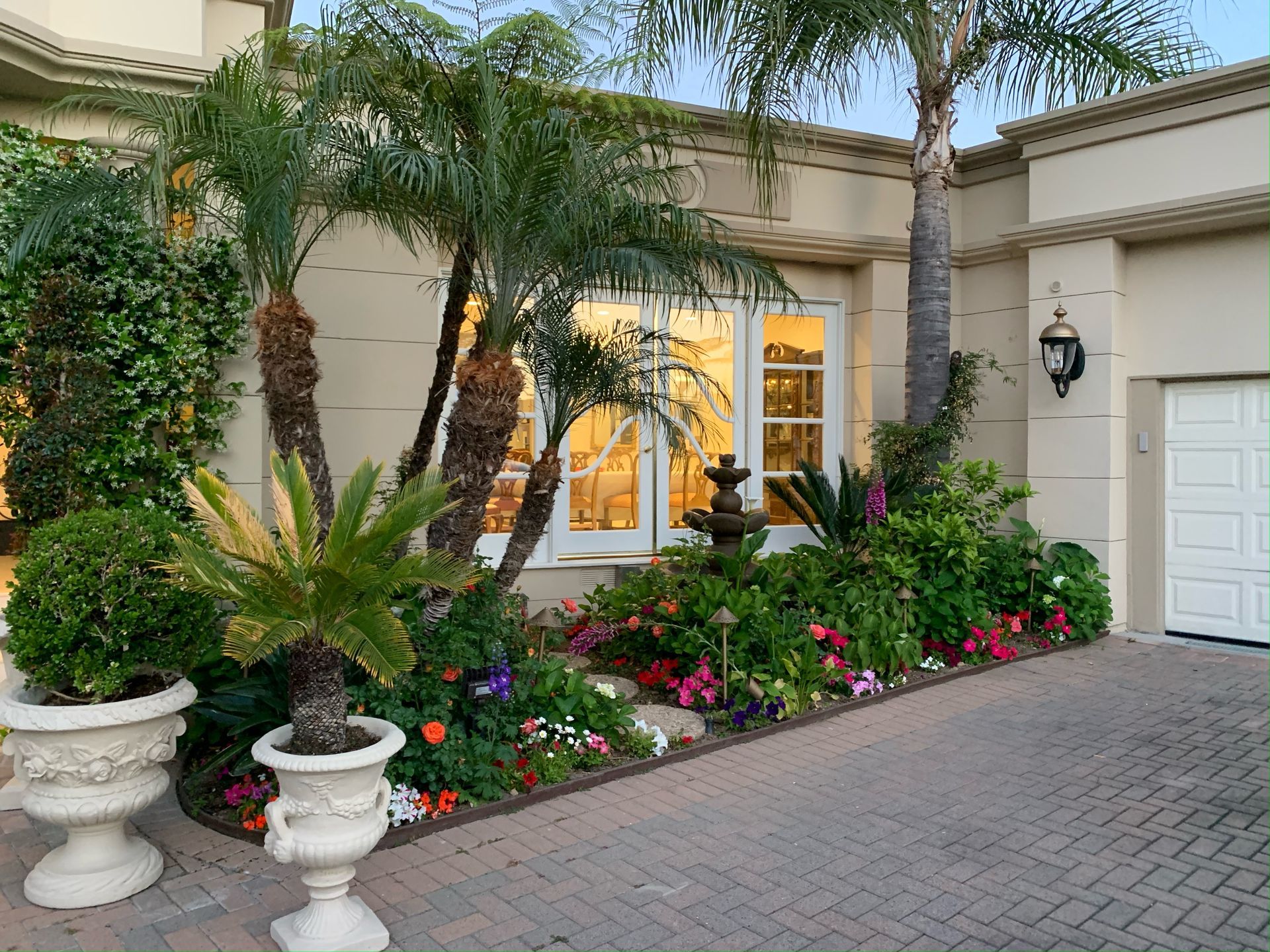A tan stucco home exterior with palm trees, a decorative fountain, potted plants, and colorful flowers in a front garden.