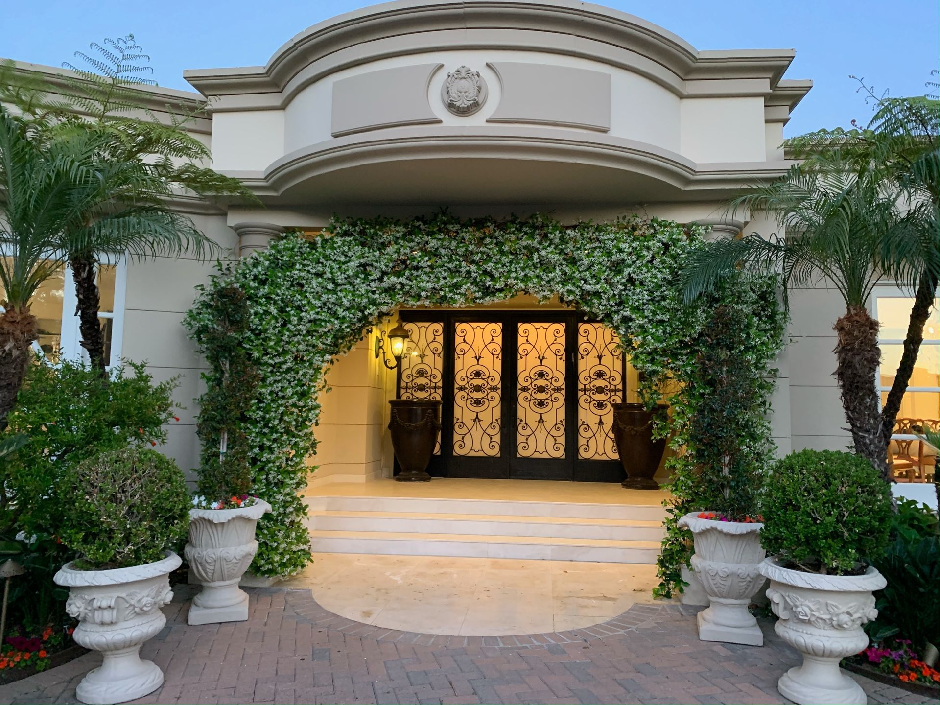 A beige building entrance framed by a lush green archway, flanked by large white decorative planters on a stone patio.