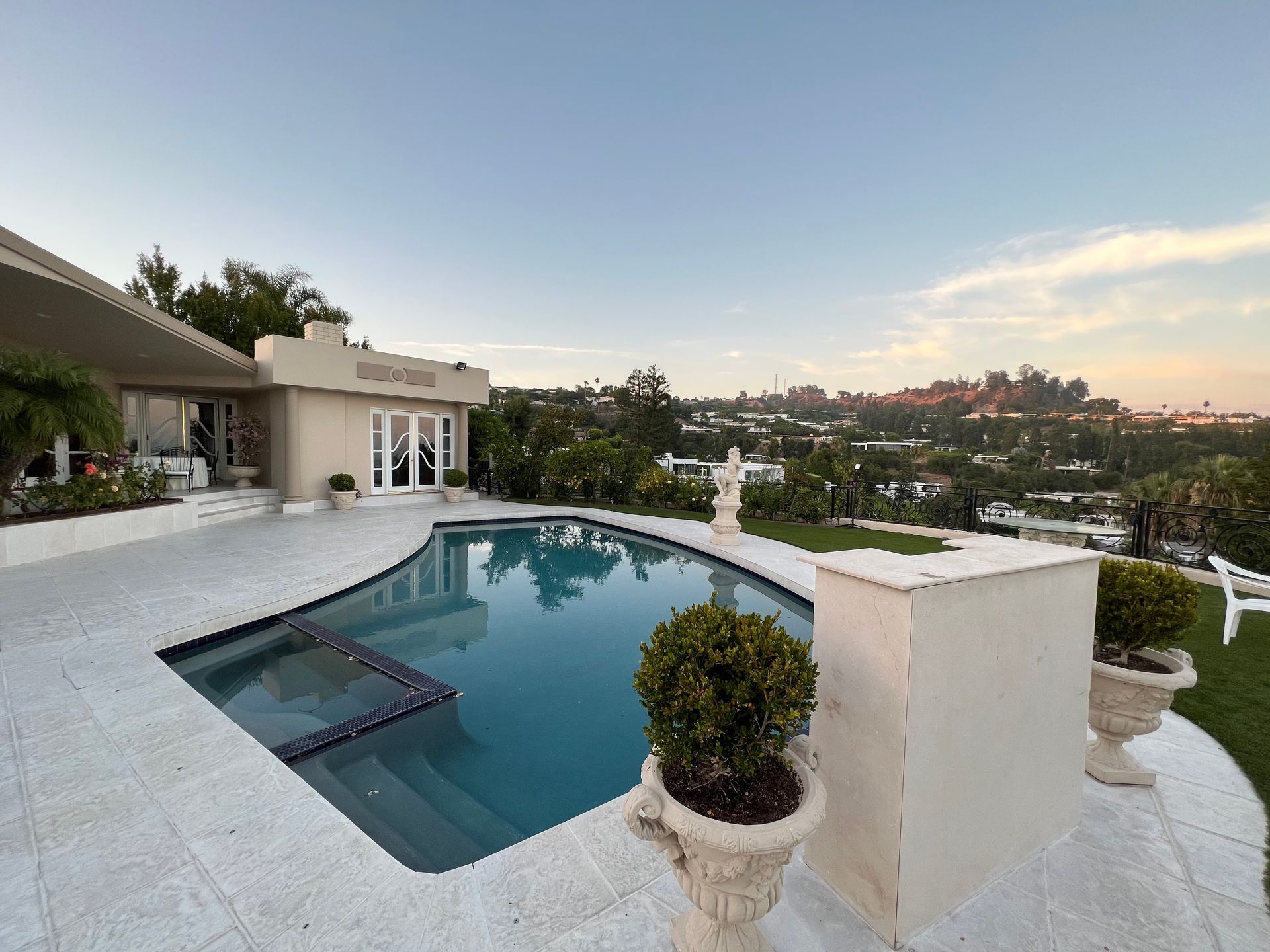 An outdoor swimming pool with a stone patio, potted plants, and a house set against a scenic hillside view at sunset.