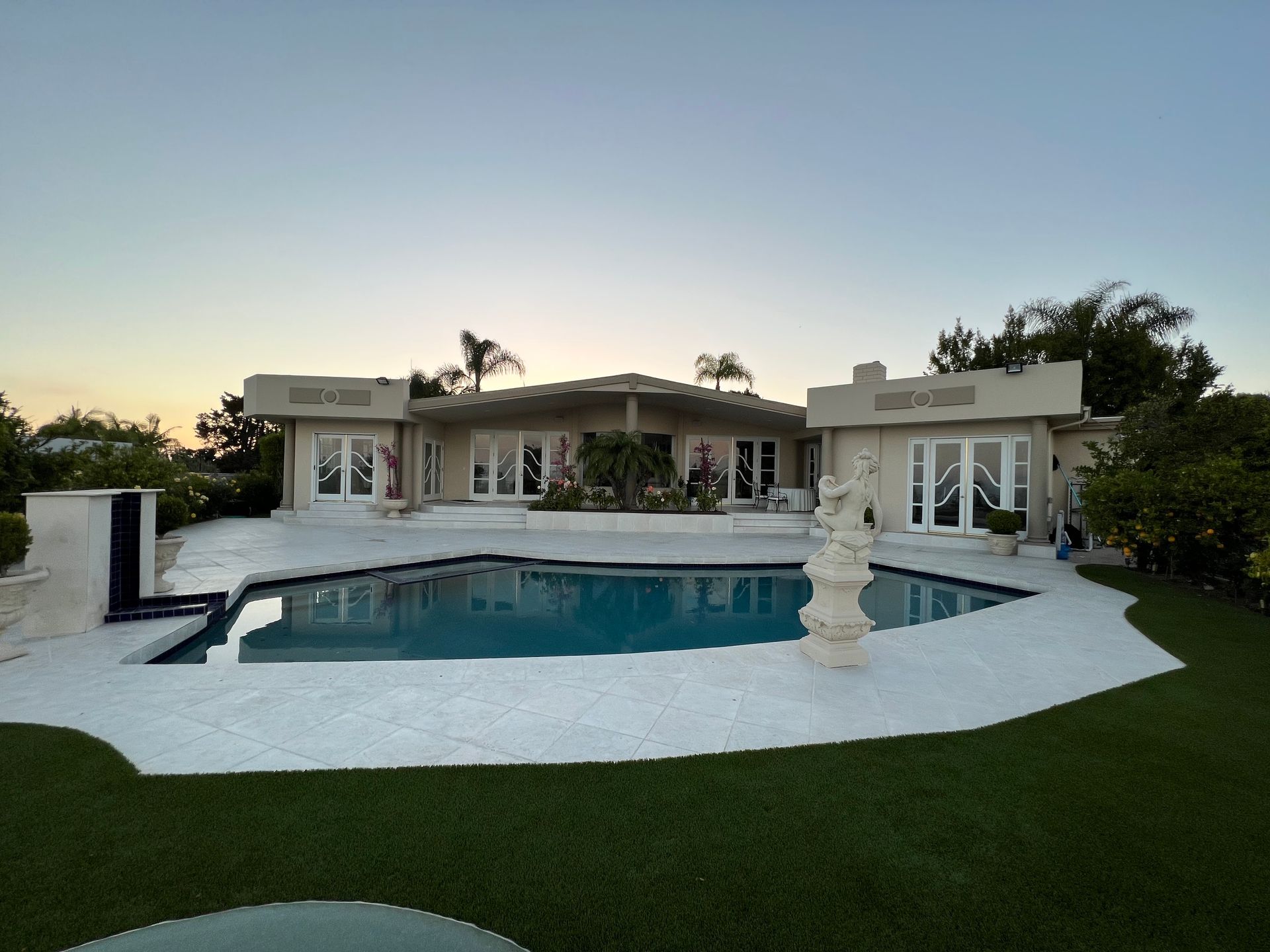 A wide shot of a beige, single-story house with a large, irregularly shaped pool and white patio under a twilight sky.
