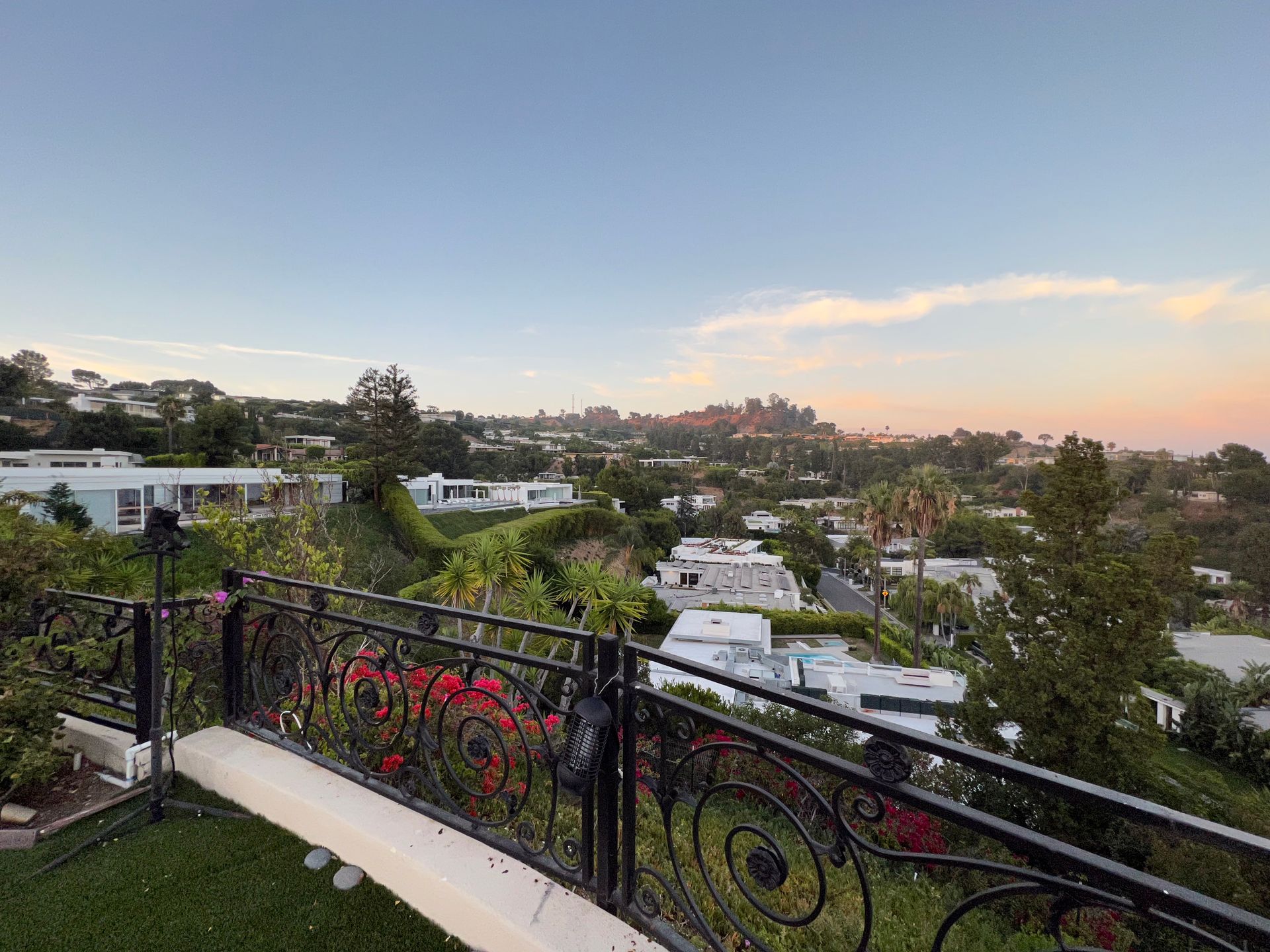 A wrought-iron balcony railing overlooks a hillside neighborhood at sunset with a view of a distant, hazy city skyline.