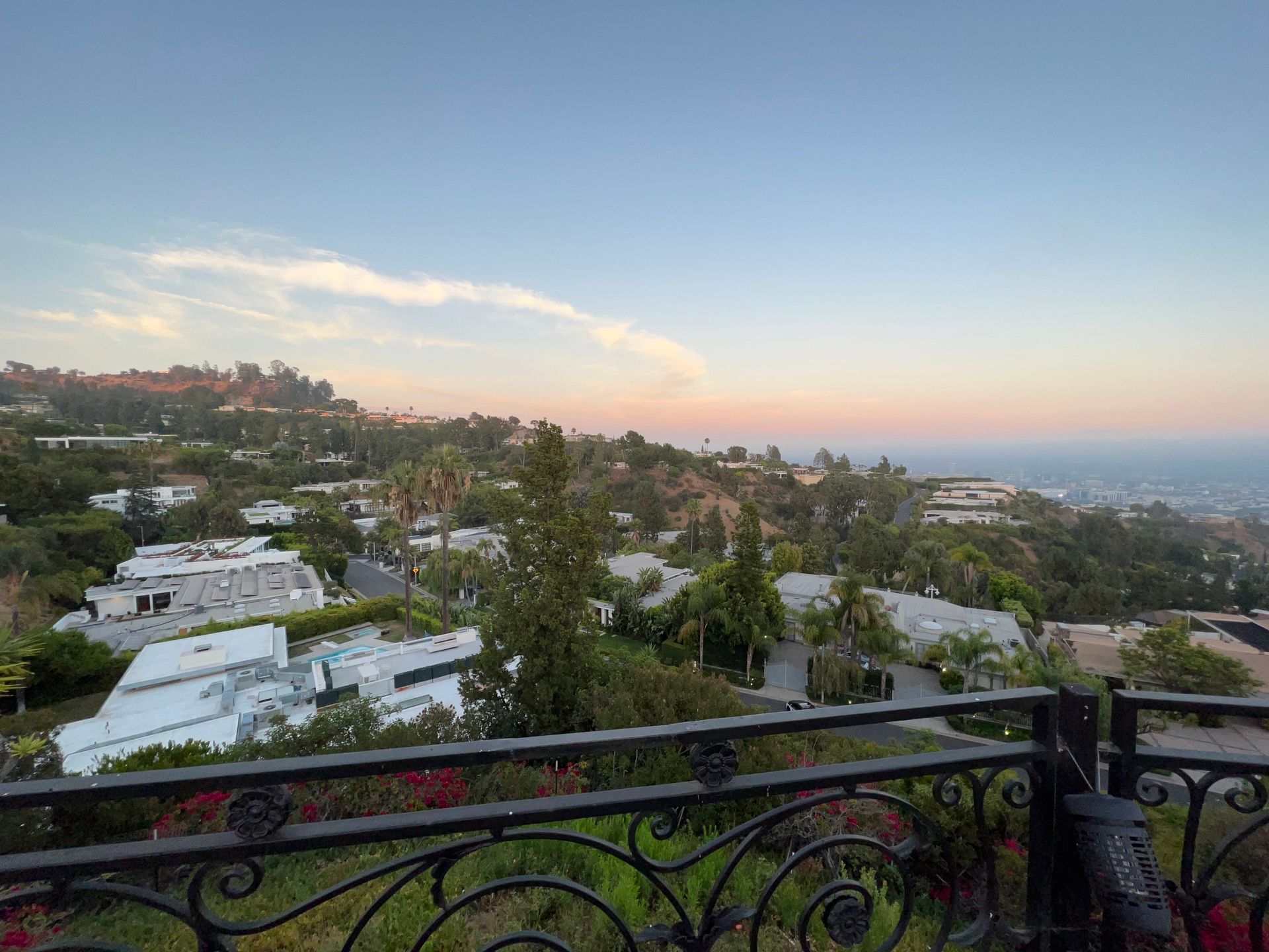 A wrought-iron balcony overlooks a hillside neighborhood with white houses toward a distant ocean at sunset.