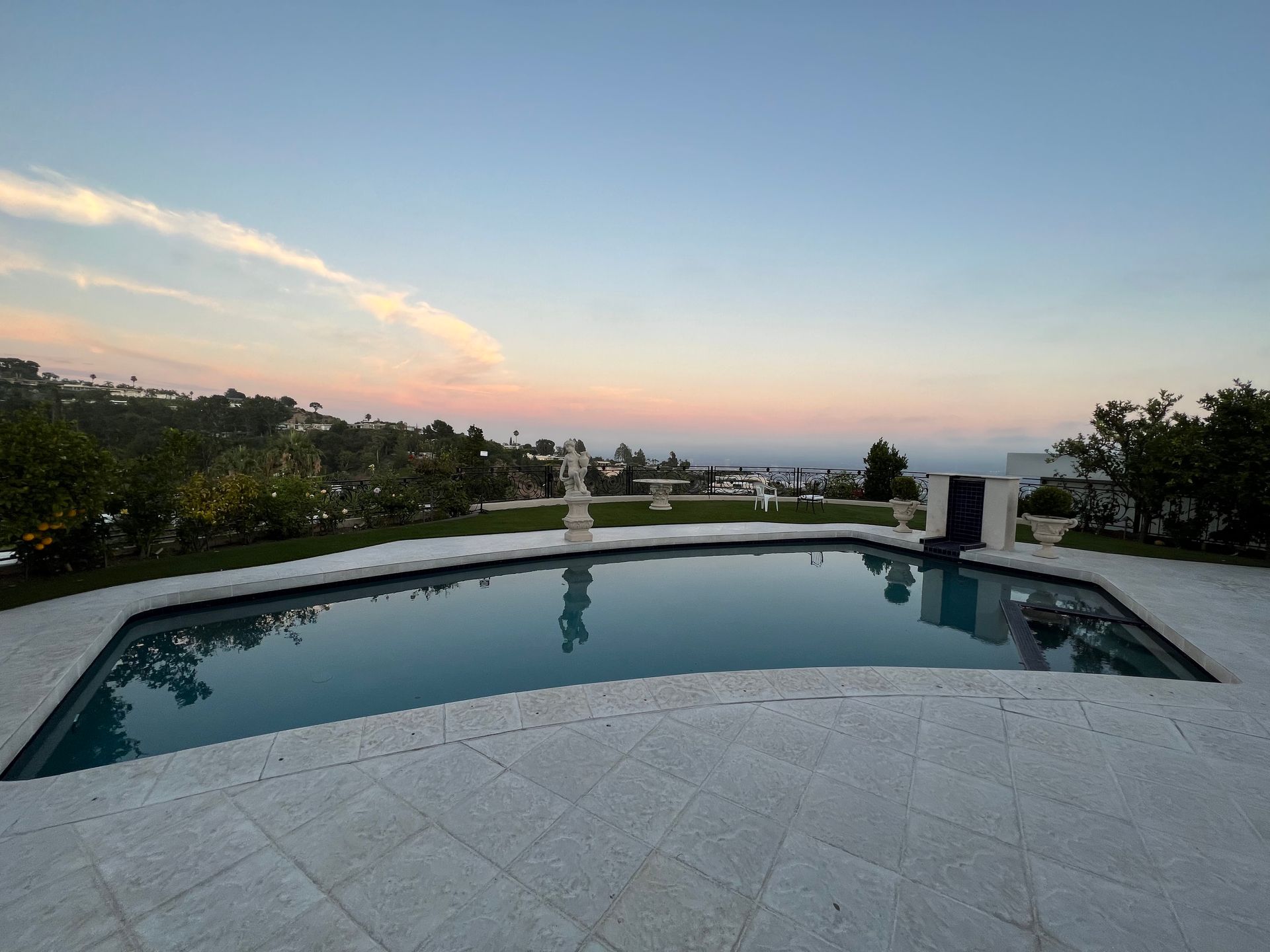A swimming pool surrounded by a stone patio overlooks a hillside landscape during a pink and orange sunset.