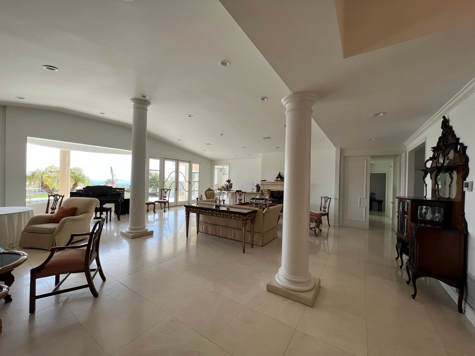 A spacious, bright open-plan living room with white tiled floors, two large decorative pillars, and a distant ocean view.