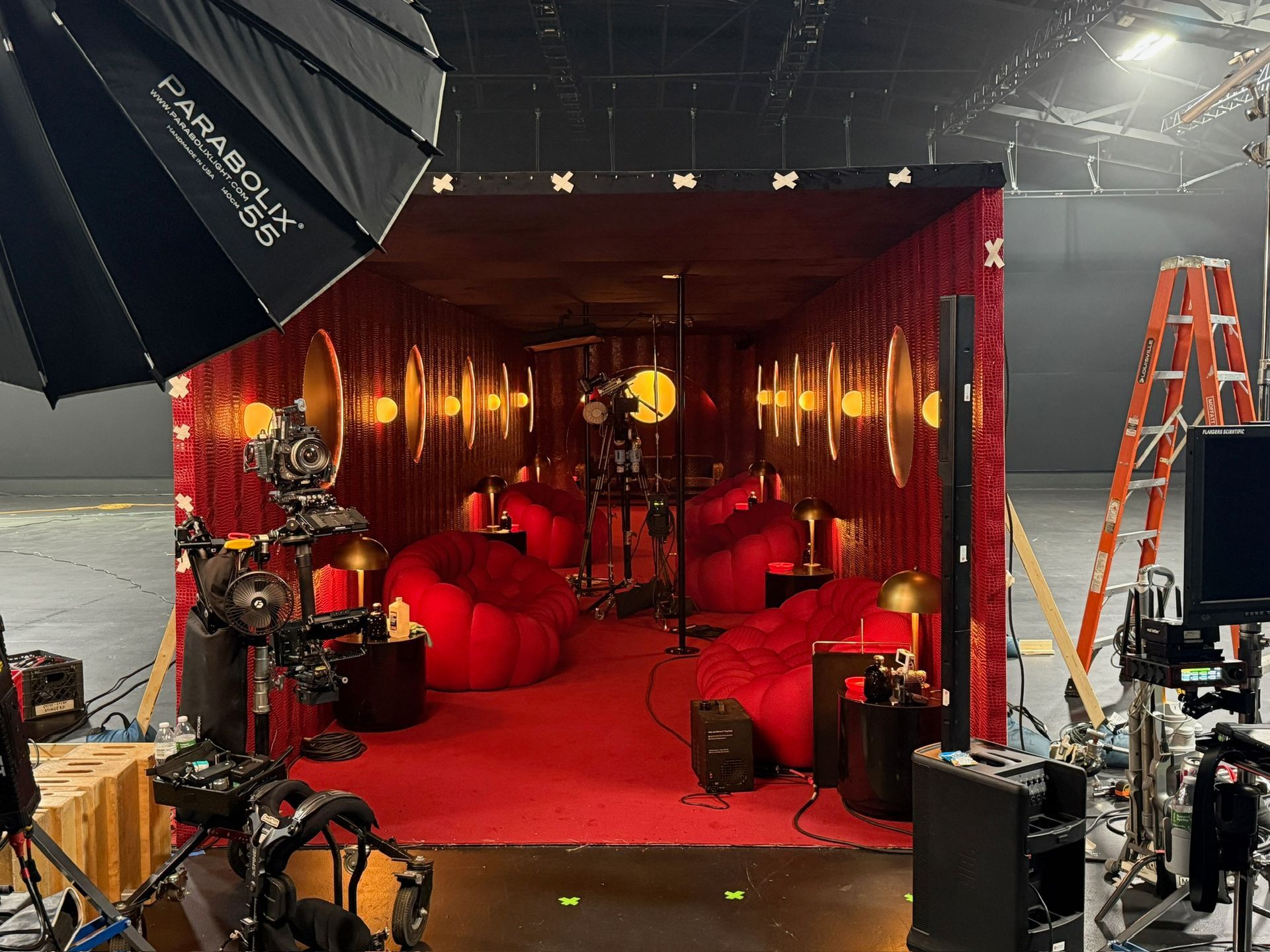 A film set interior features a red room with round chairs, golden lighting, and professional video production equipment.