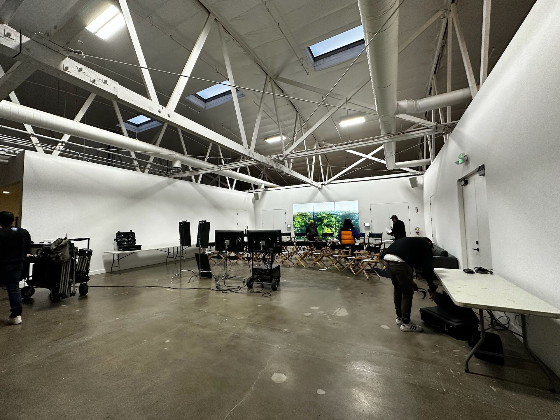A studio with high ceilings, exposed trusses, and concrete floors, showing people working near screens and equipment.