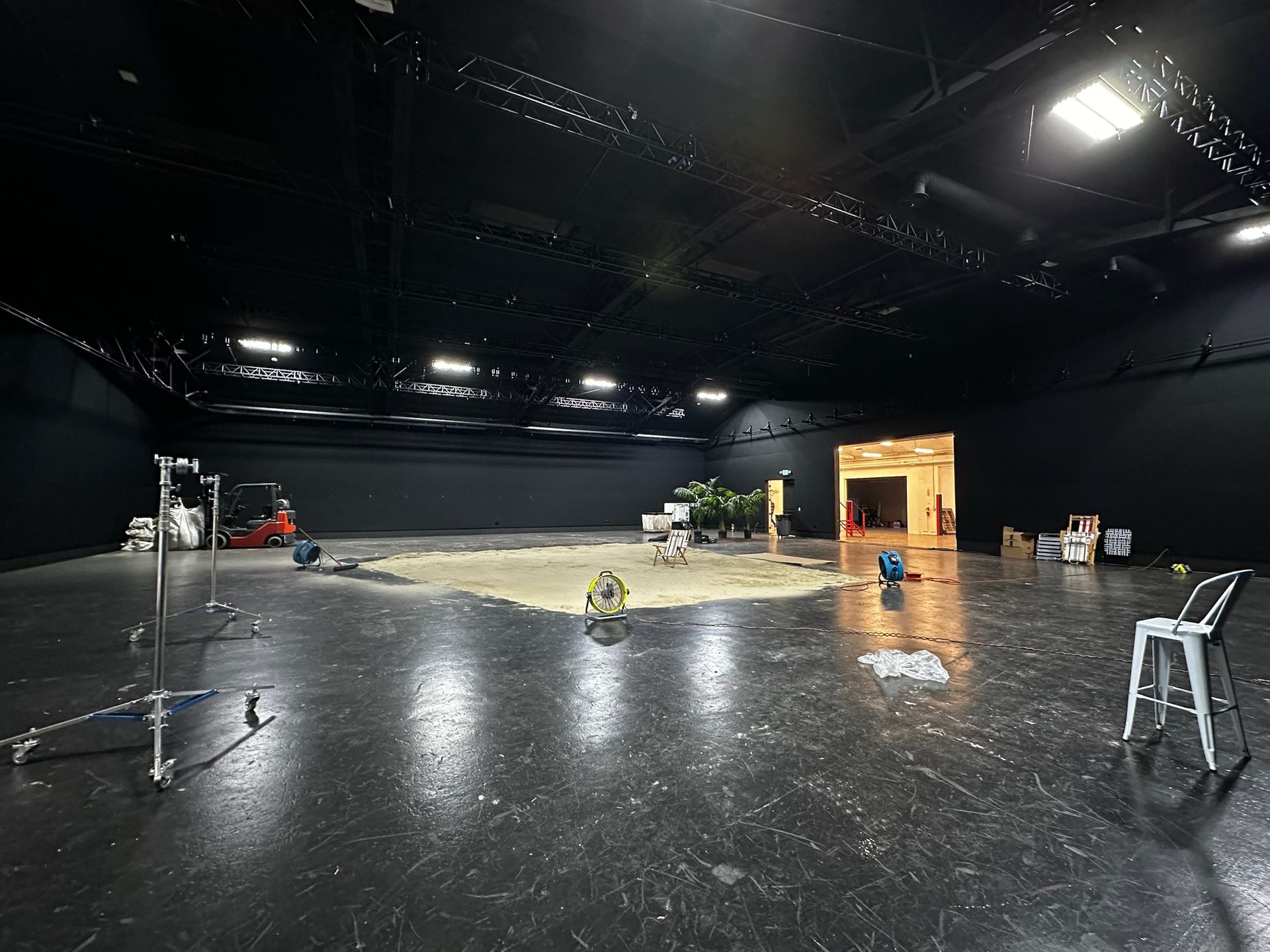 A wide shot of a large, empty black studio with a patch of sand in the center, lighting equipment, and a white chair.