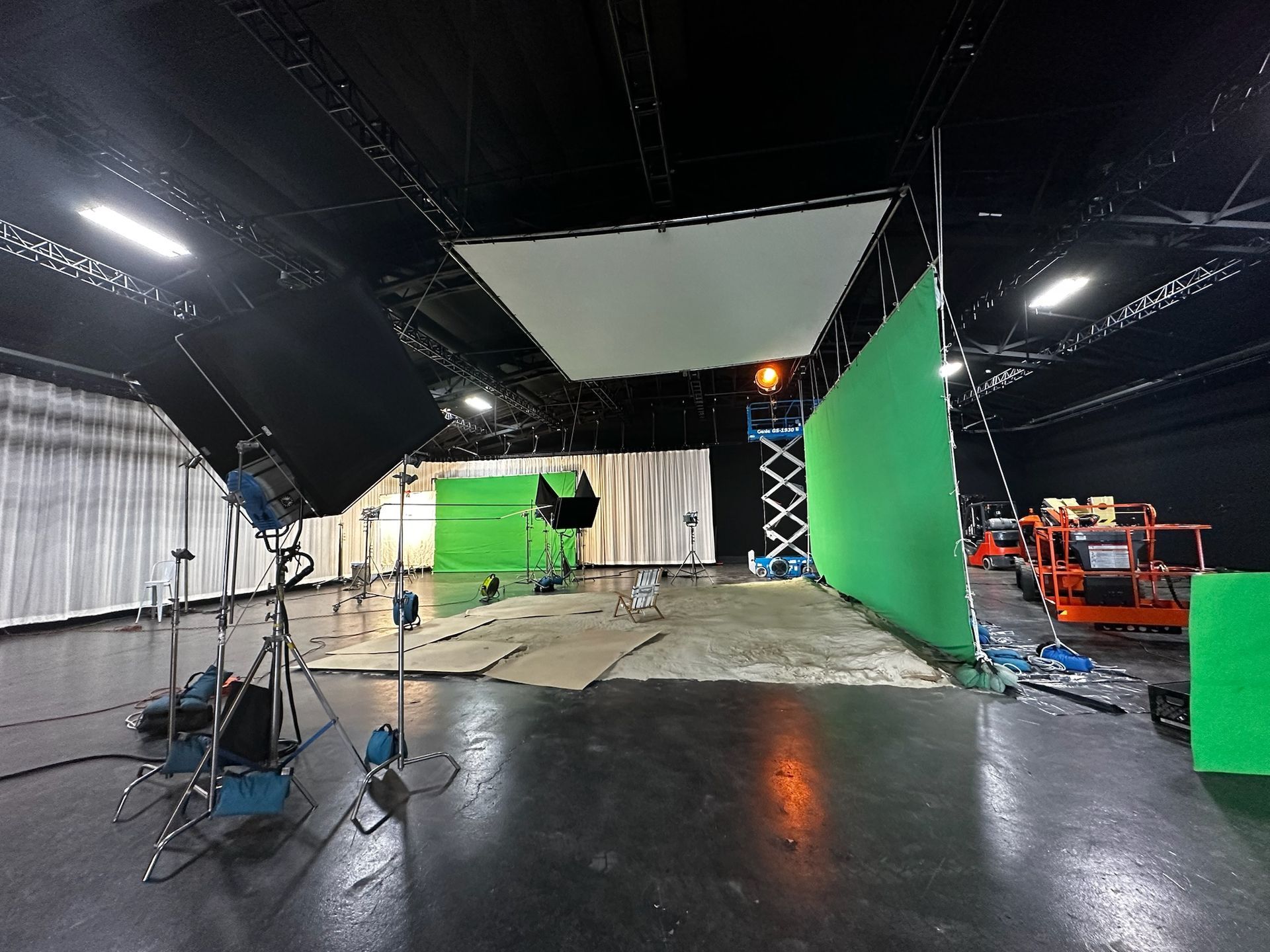 A film studio with a large green screen, overhead lighting panels, and professional production equipment on a dark floor.