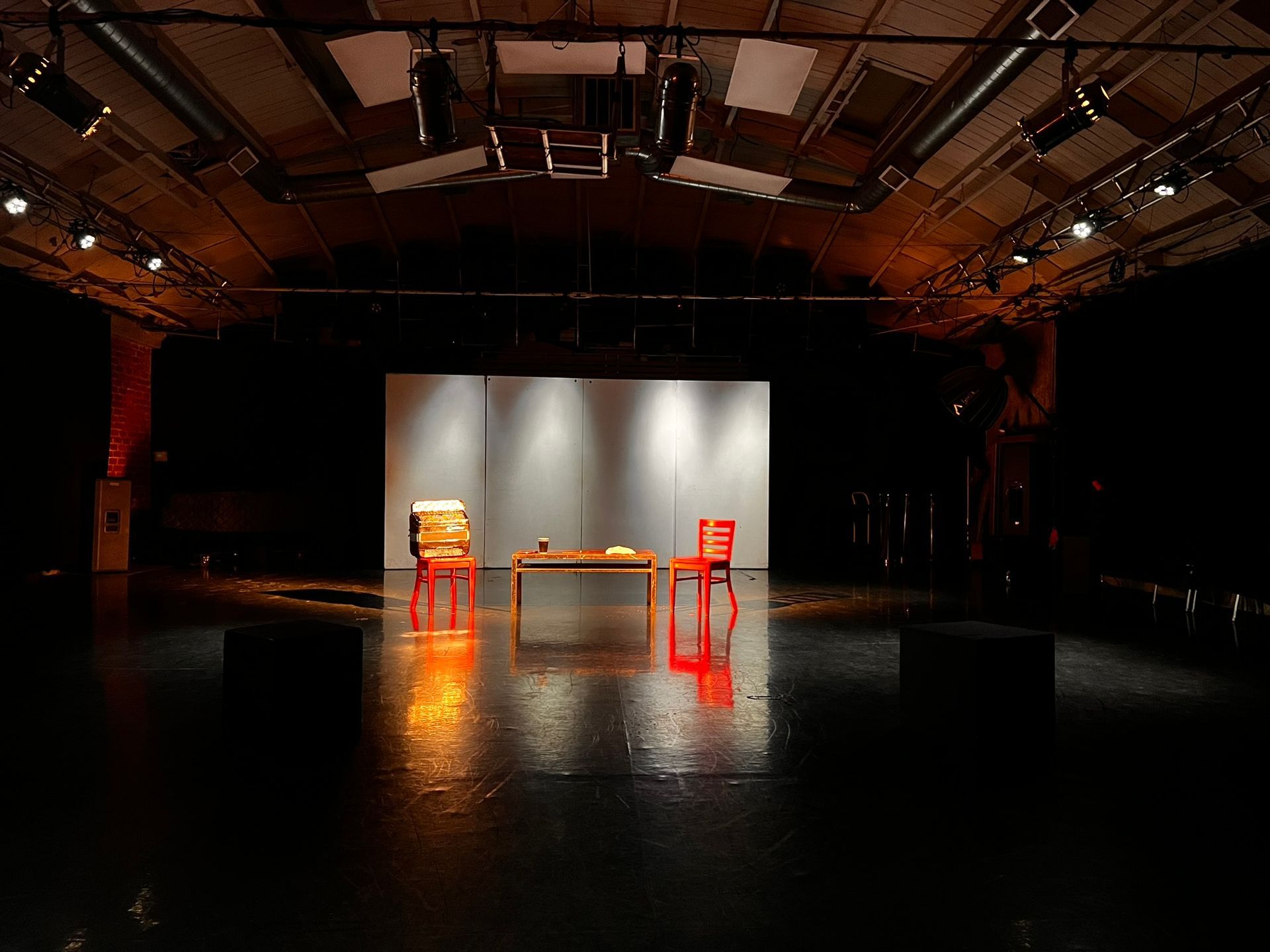 A stage featuring a wooden table flanked by two orange chairs, illuminated by overhead stage lights against a grey backdrop.
