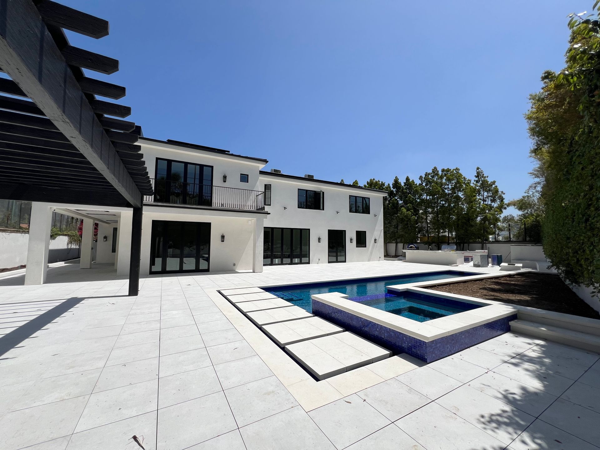 A white two-story house with a black pergola, a swimming pool, and a white tiled patio under a clear blue sky.