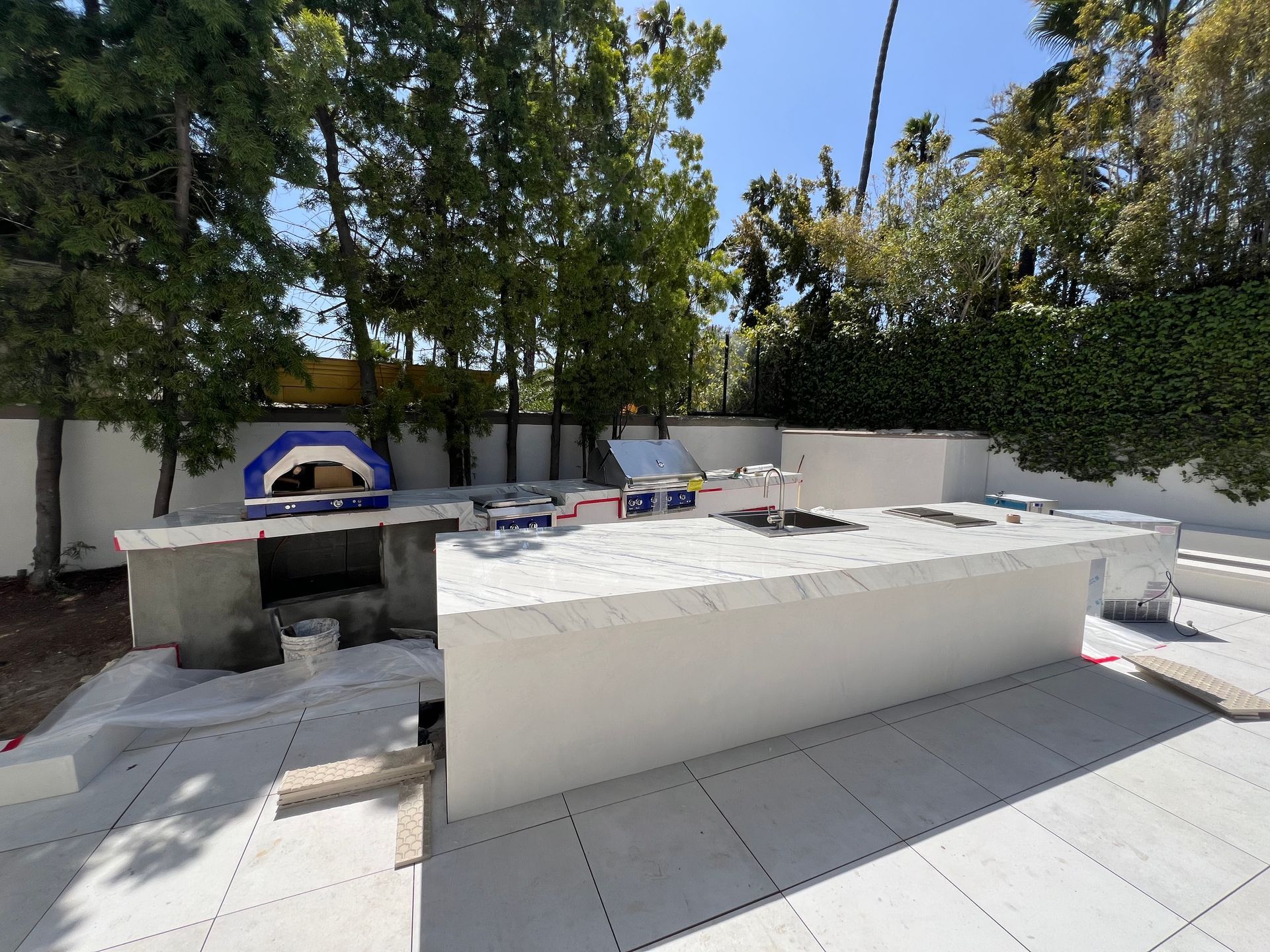 An outdoor kitchen construction site with a long white marble island, a pizza oven, and paved patio flooring.