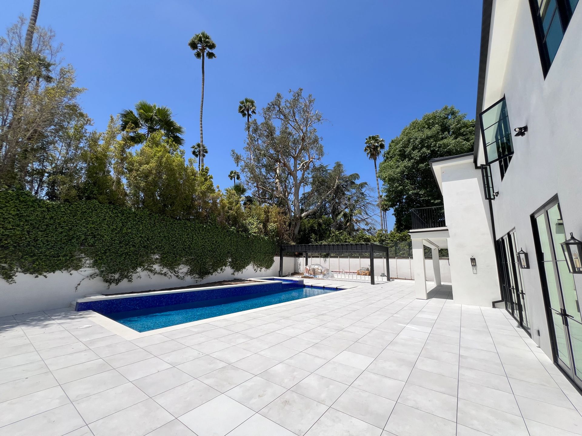 A bright white house with a patio and rectangular blue swimming pool set against a green hedge and tall palm trees.