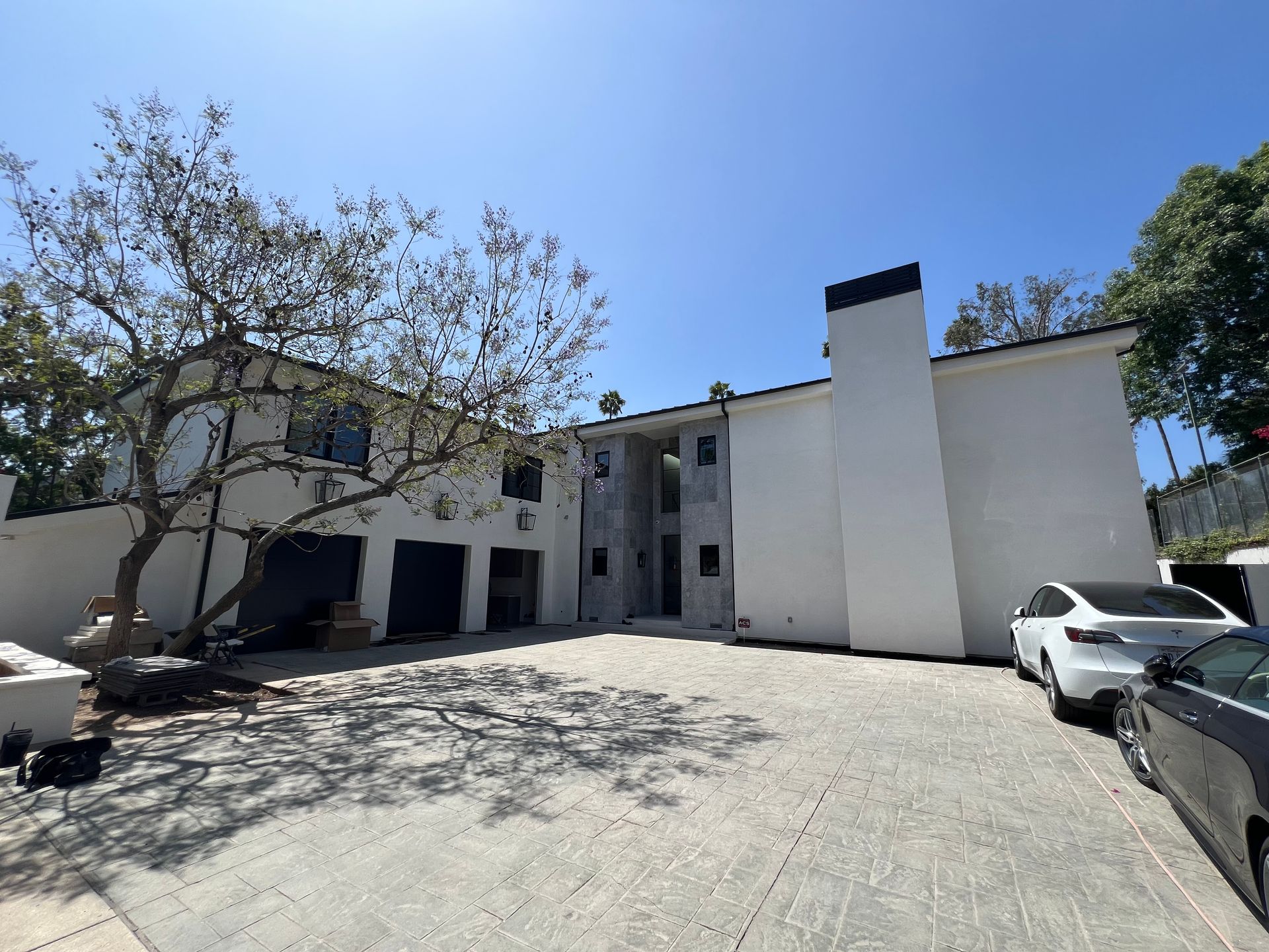 A white two-story house with a three-car garage, a tree in the yard, and a paved driveway with two parked cars.