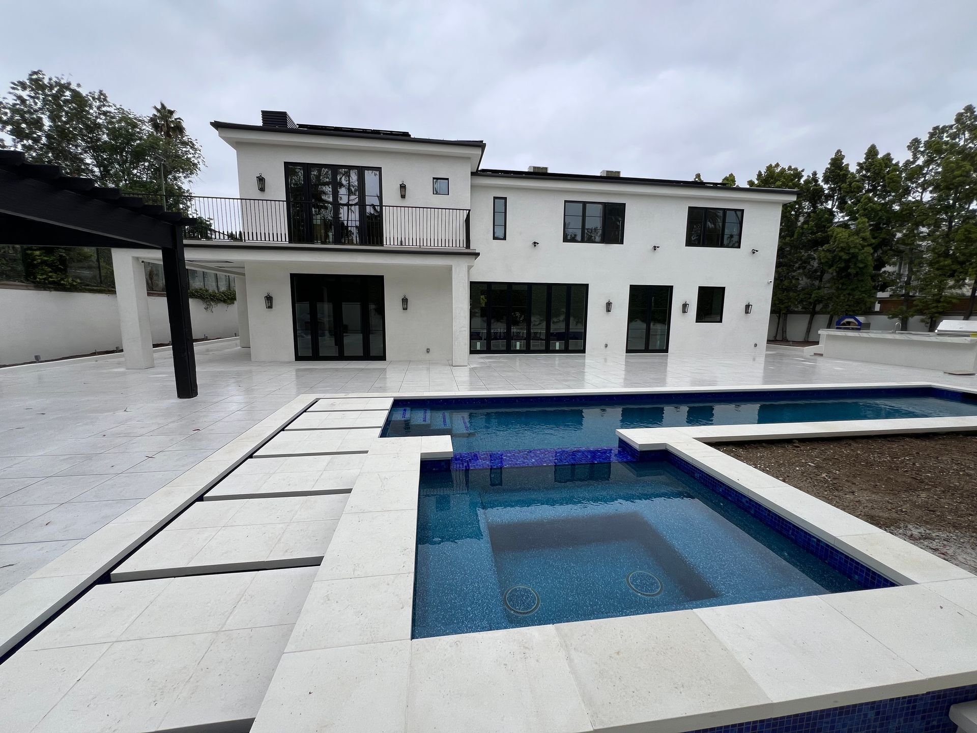 A white two-story house with a modern pool and spa featuring blue tiled interior and light-colored stone decking.