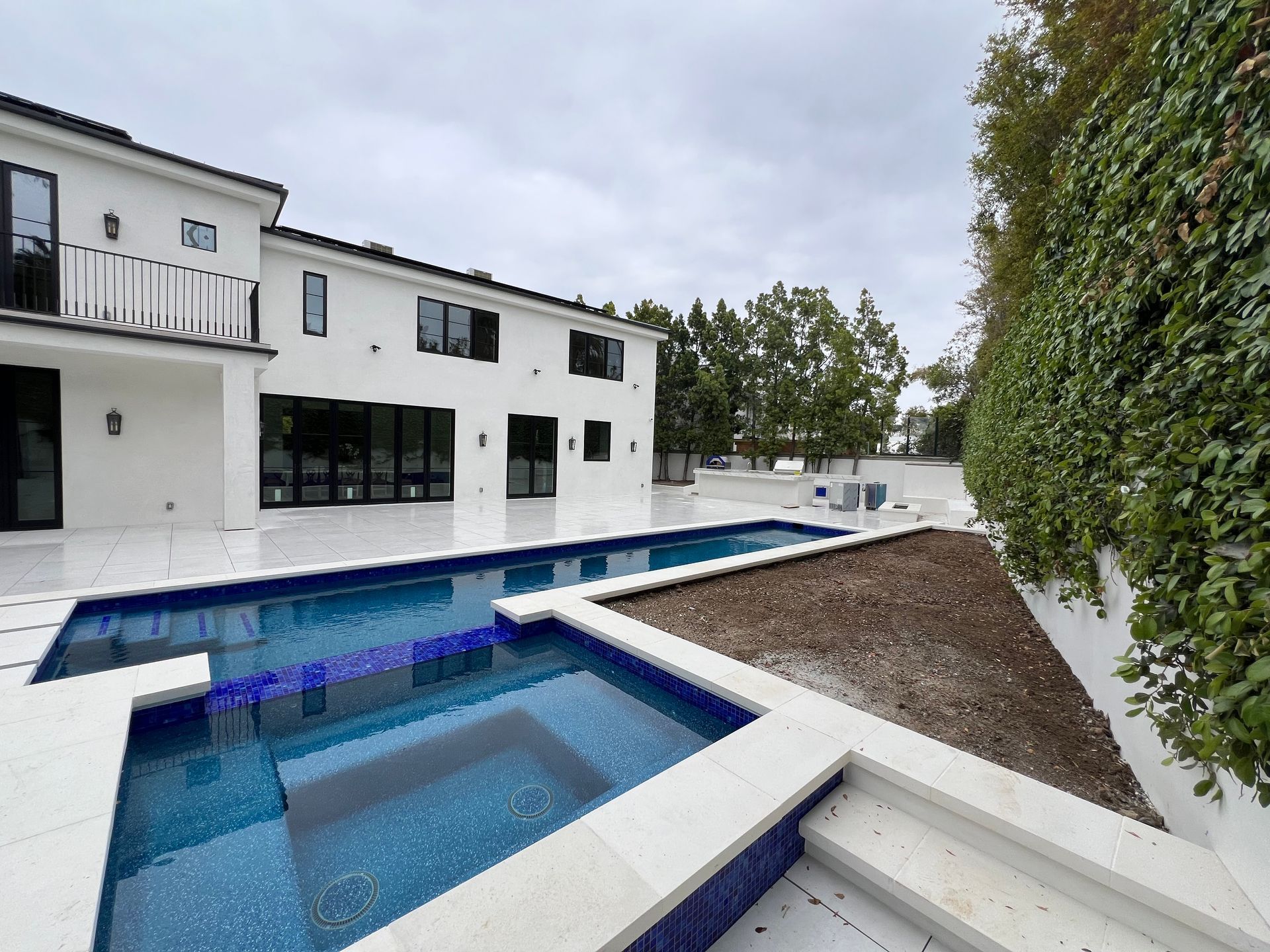 A modern white house with a blue swimming pool, a square hot tub, and a tall ivy-covered wall.