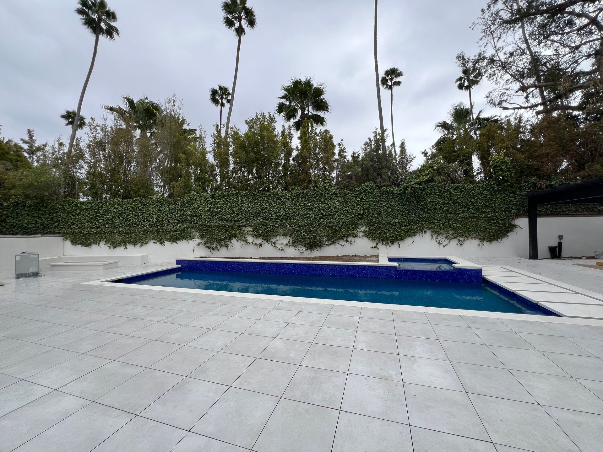 A rectangular blue swimming pool set in a large, light-colored stone patio surrounded by a wall of vines and palm trees.
