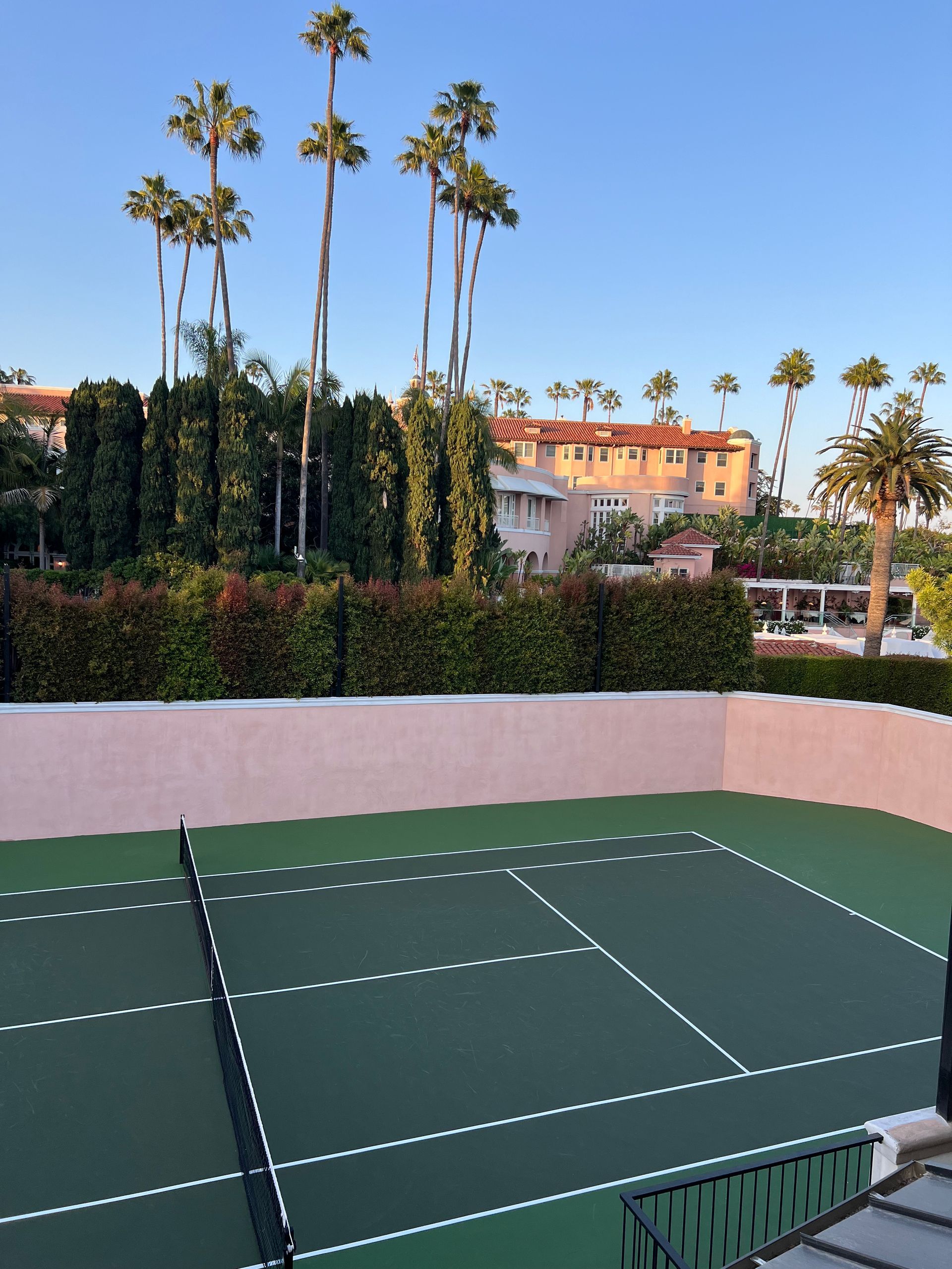 A high-angle view of a green pickleball court bordered by tall hedges, palm trees, and a Spanish-style building.