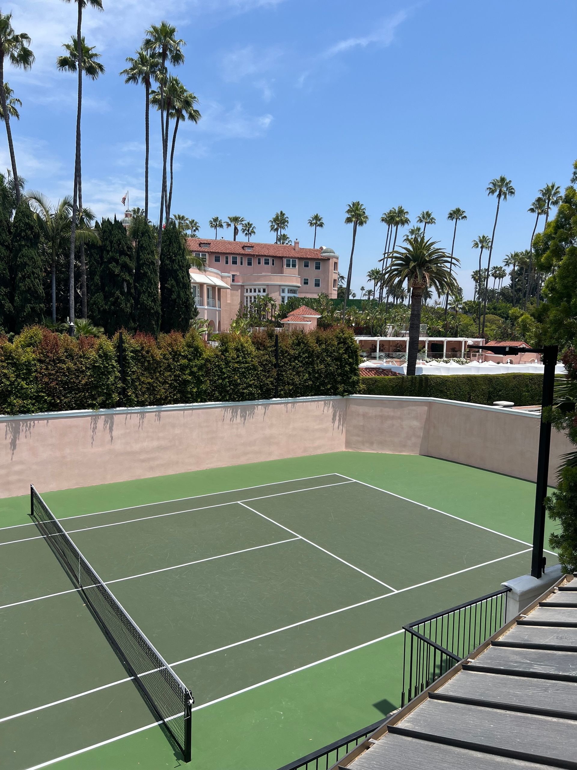An outdoor tennis court with a green surface and white lines, bordered by a beige wall and palm trees under a blue sky.