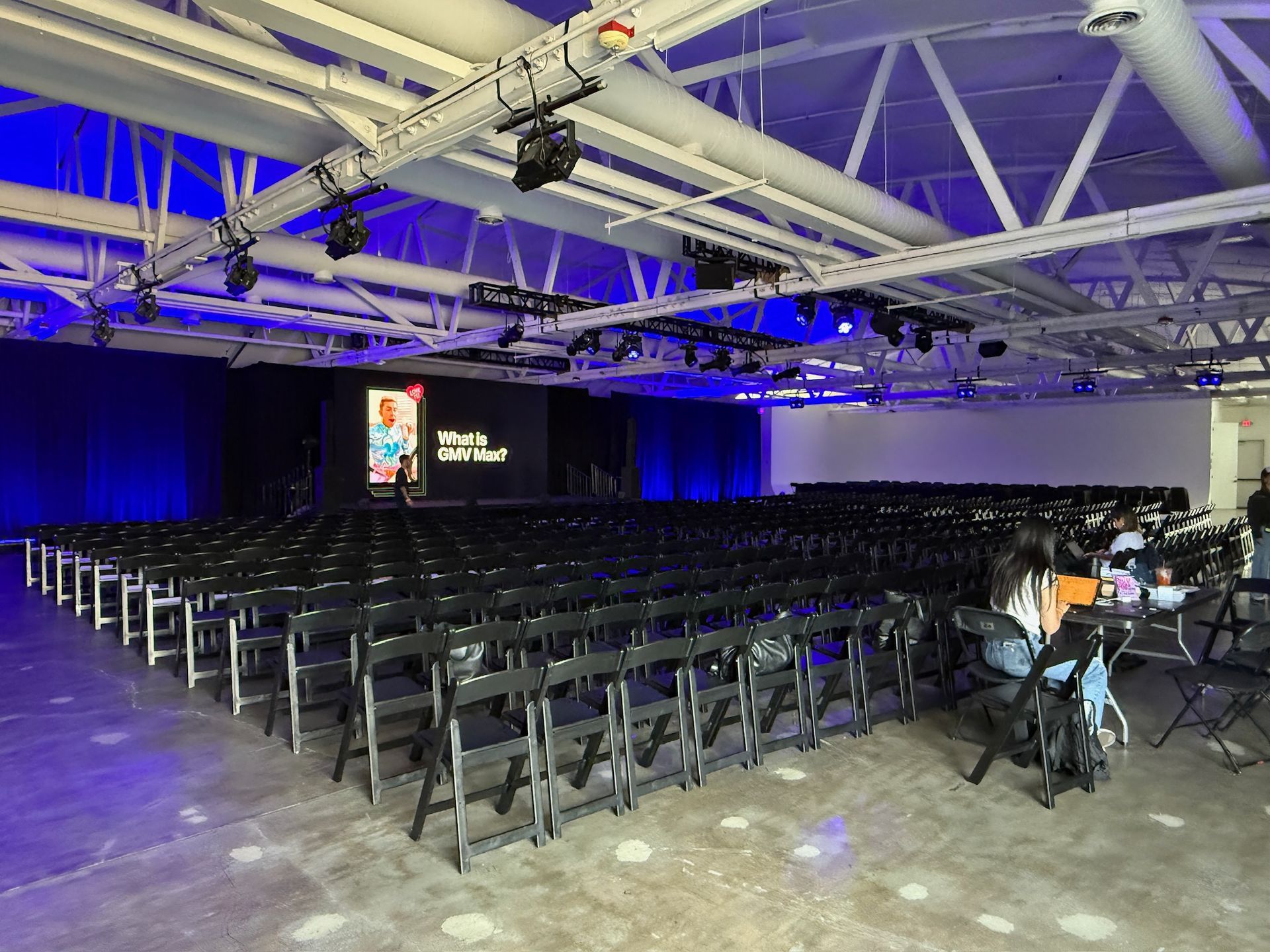 An auditorium with rows of black chairs facing a stage with a digital display, illuminated by blue overhead lighting.