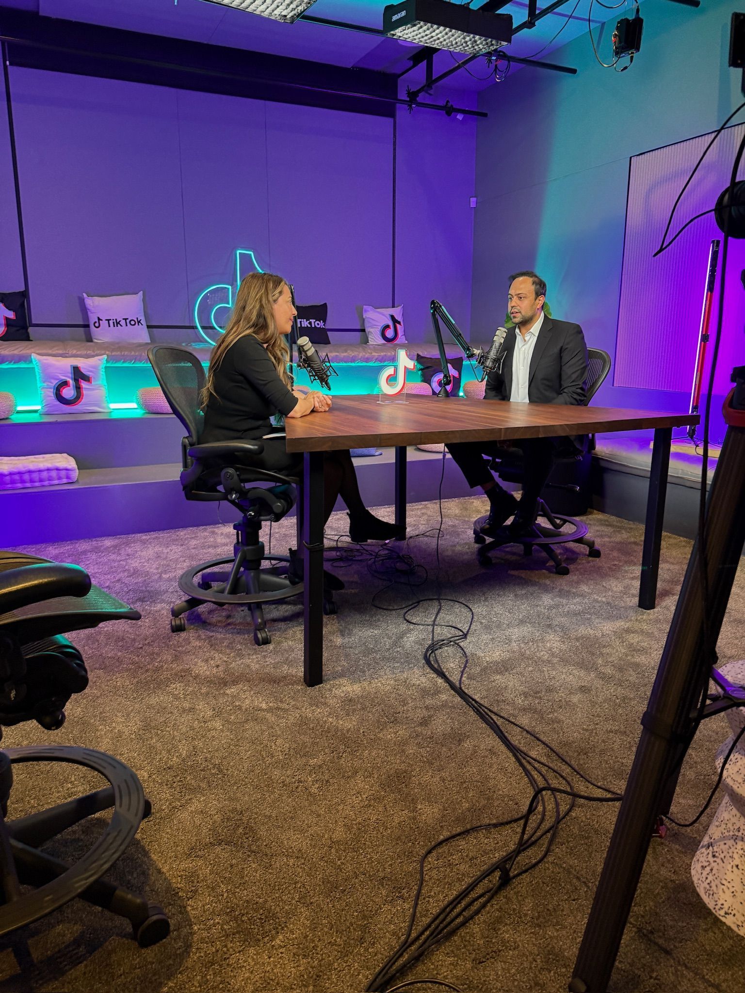 Two people sit opposite each other at a wooden table in a purple-lit studio with microphones, recording a podcast.