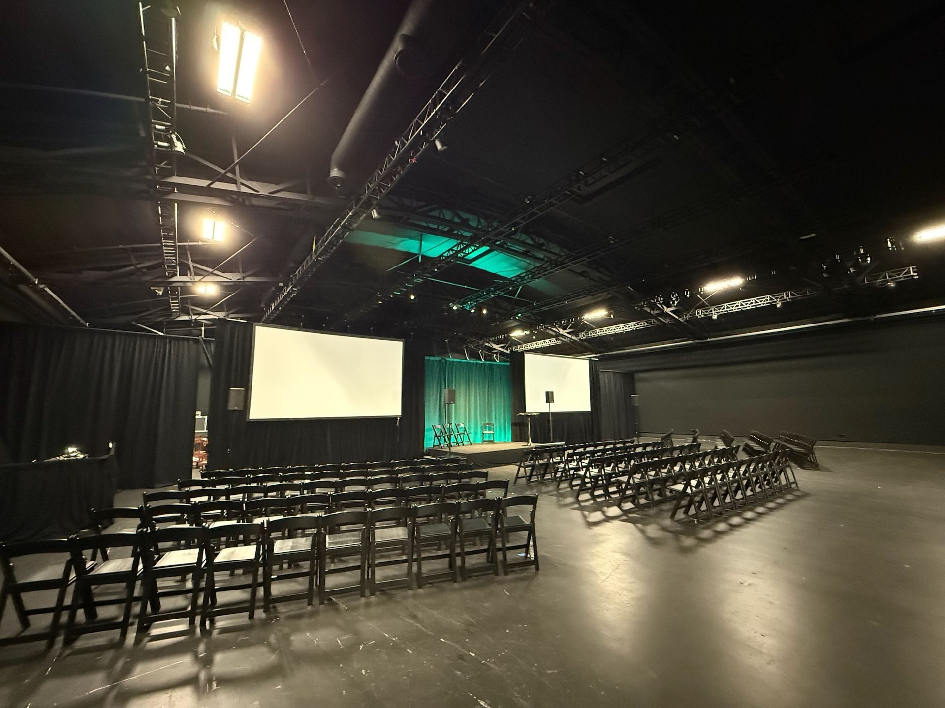 An empty event space with rows of folding chairs, two large projection screens, and stage lighting.