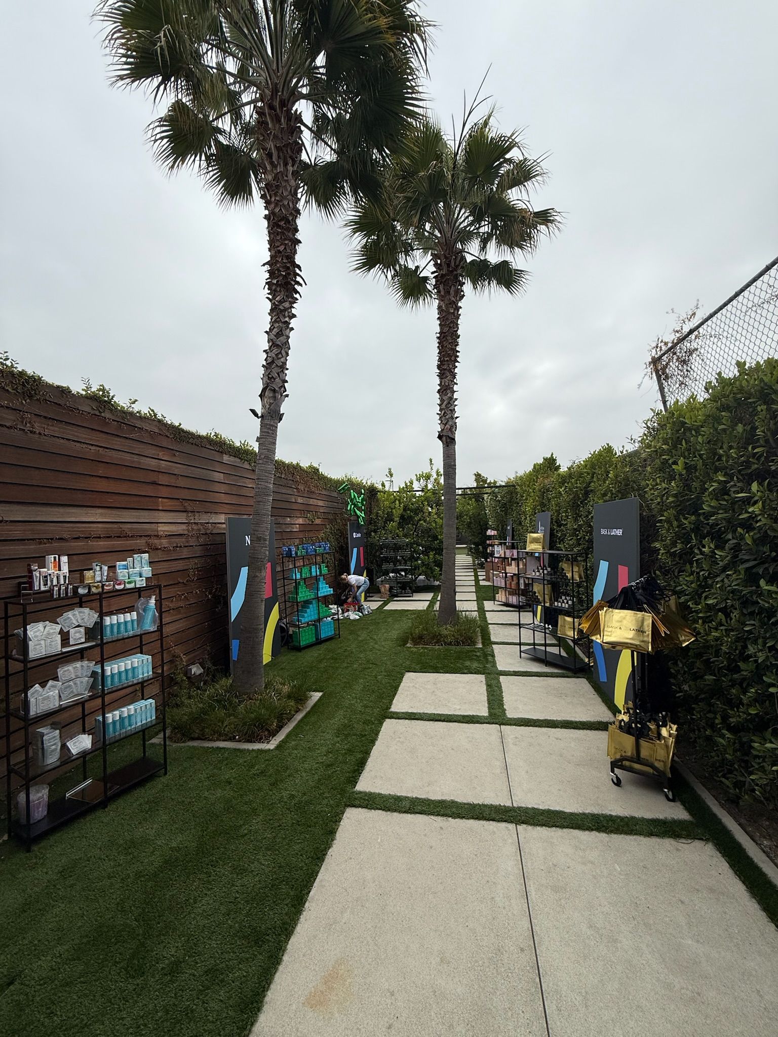A path paved with rectangular stones winds through a garden with two tall palm trees, flanked by product display racks.