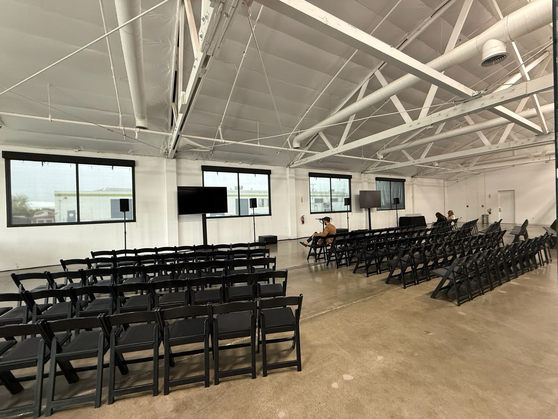 A large, white, industrial event space with rows of black folding chairs facing a screen on a stand.