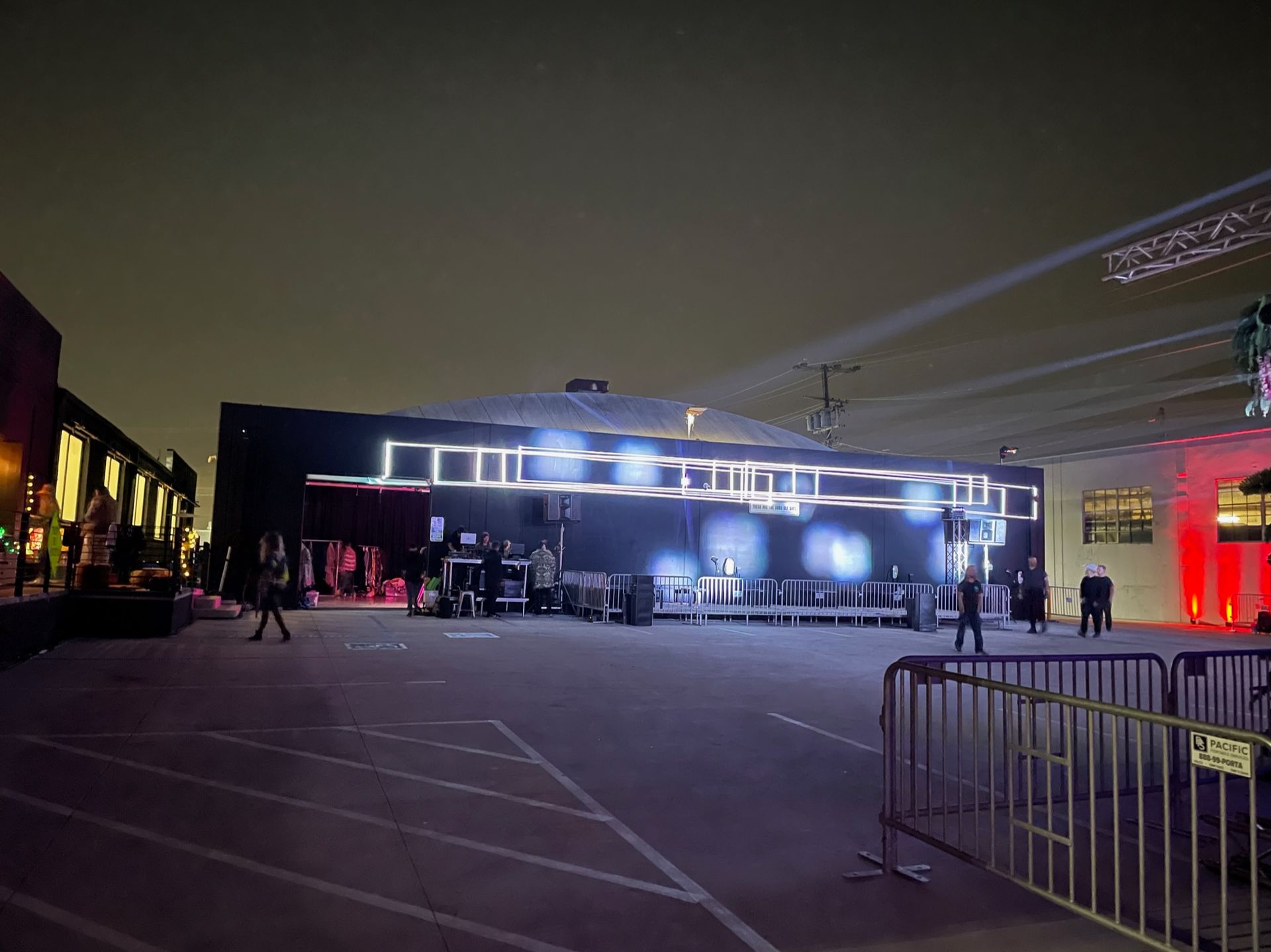 An outdoor, nighttime concert venue setup with a large dark tent, illuminated white neon accents, and metal crowd barriers.