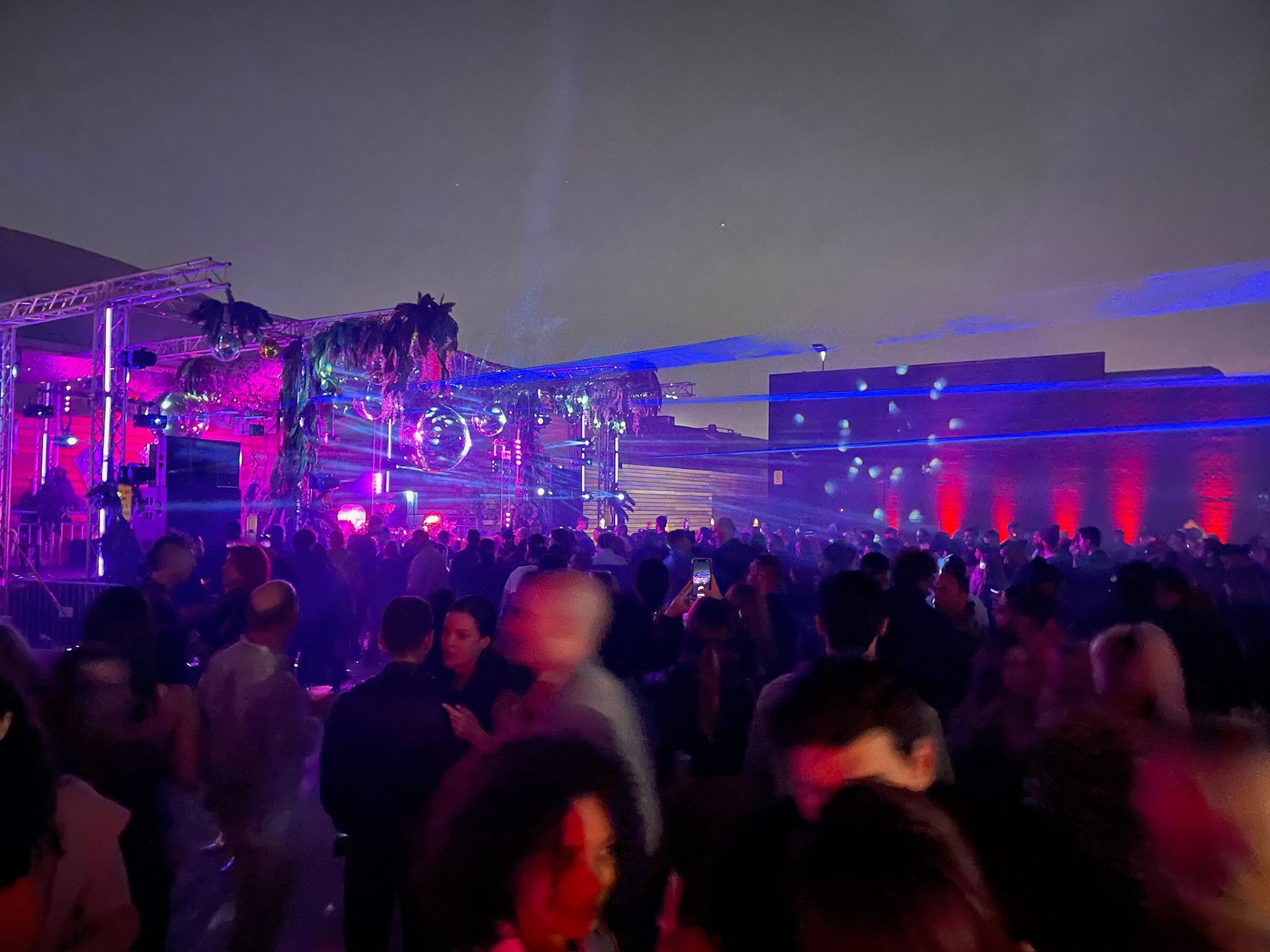 A crowded outdoor evening party with vibrant pink and blue laser lights illuminating the people and the night sky.
