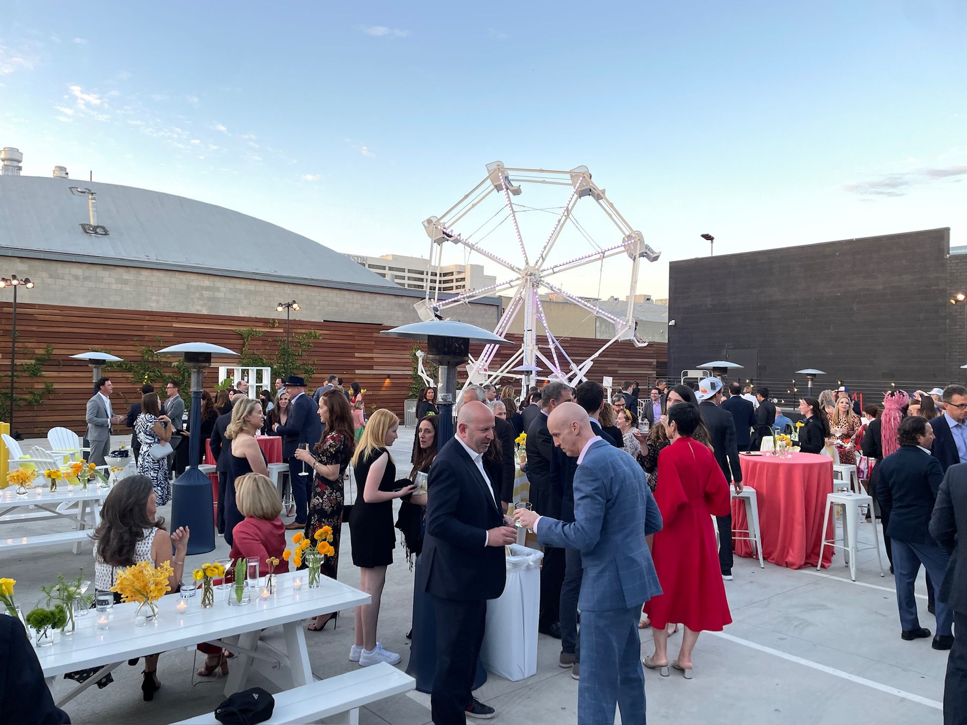 People attend an outdoor reception on a patio with tables, patio heaters, and a Ferris wheel in the background.