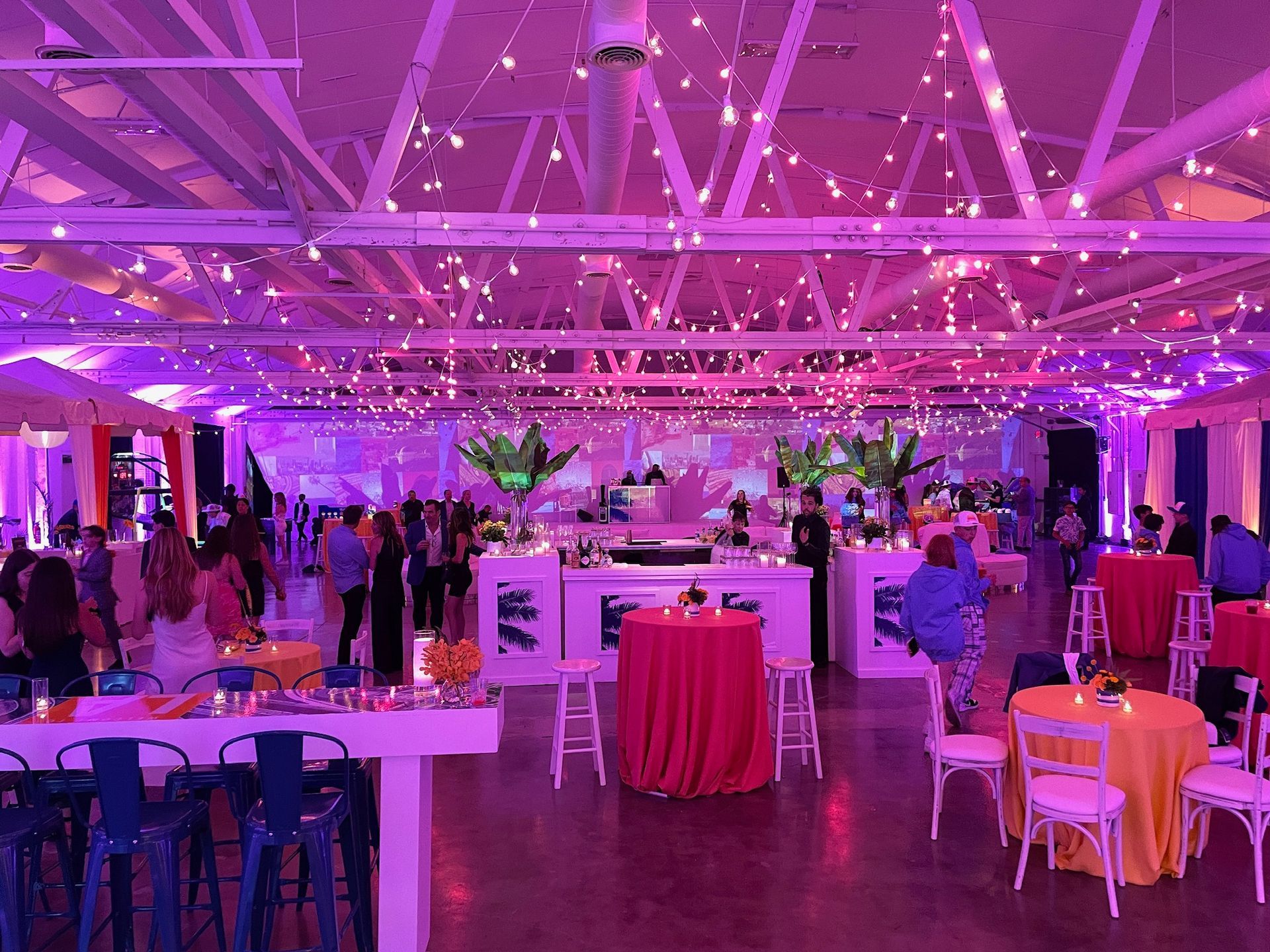 A dimly lit event space with purple lighting, string lights, a bar, and round tables with orange tablecloths.